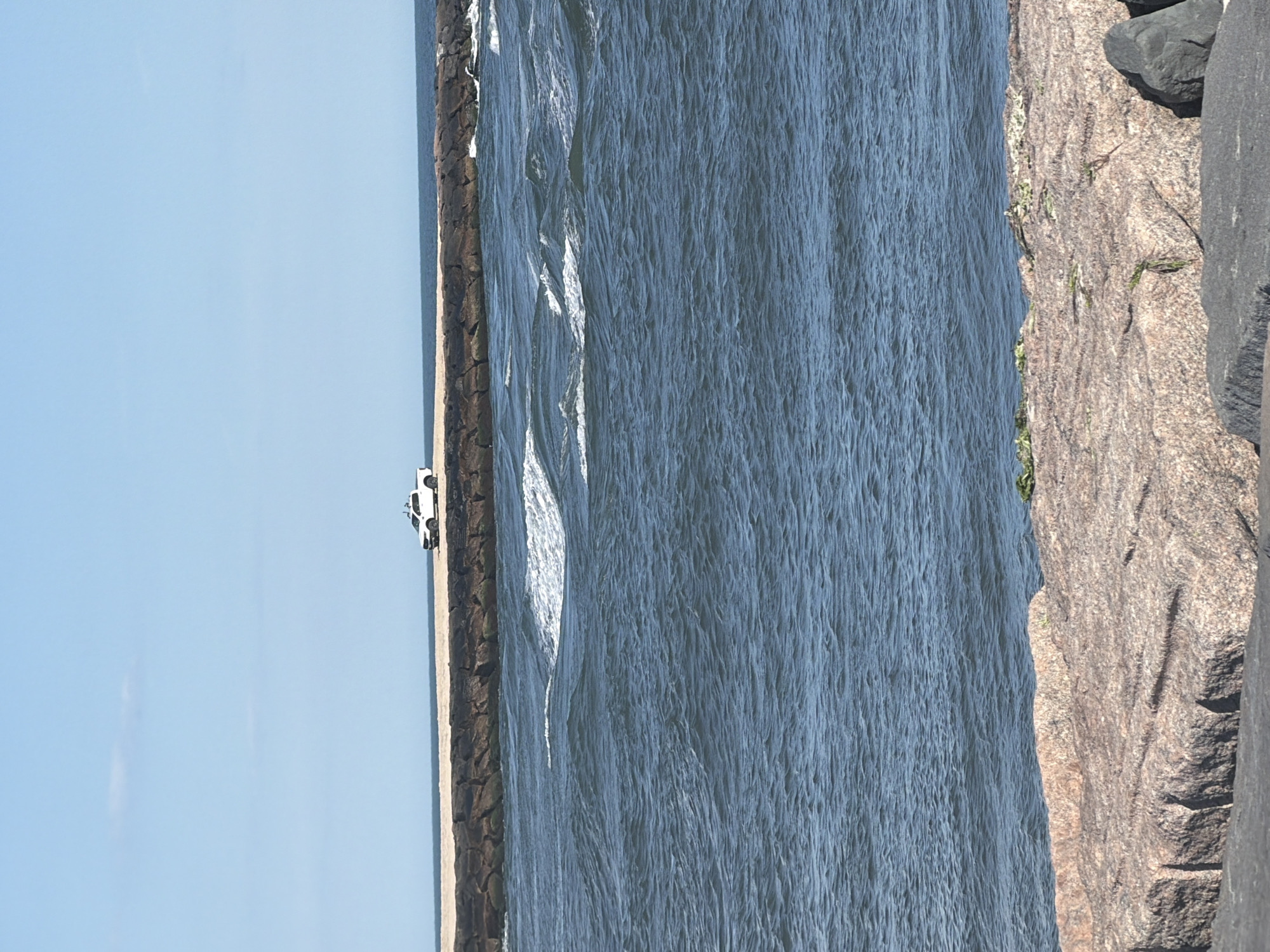 A white pickup truck is parked on a causeway. The causeway is made of large stones and sand, with a breakwater of large stones in the foreground. The ocean is visible in the background, with waves crashing against the breakwater. The sky is clear and blue.