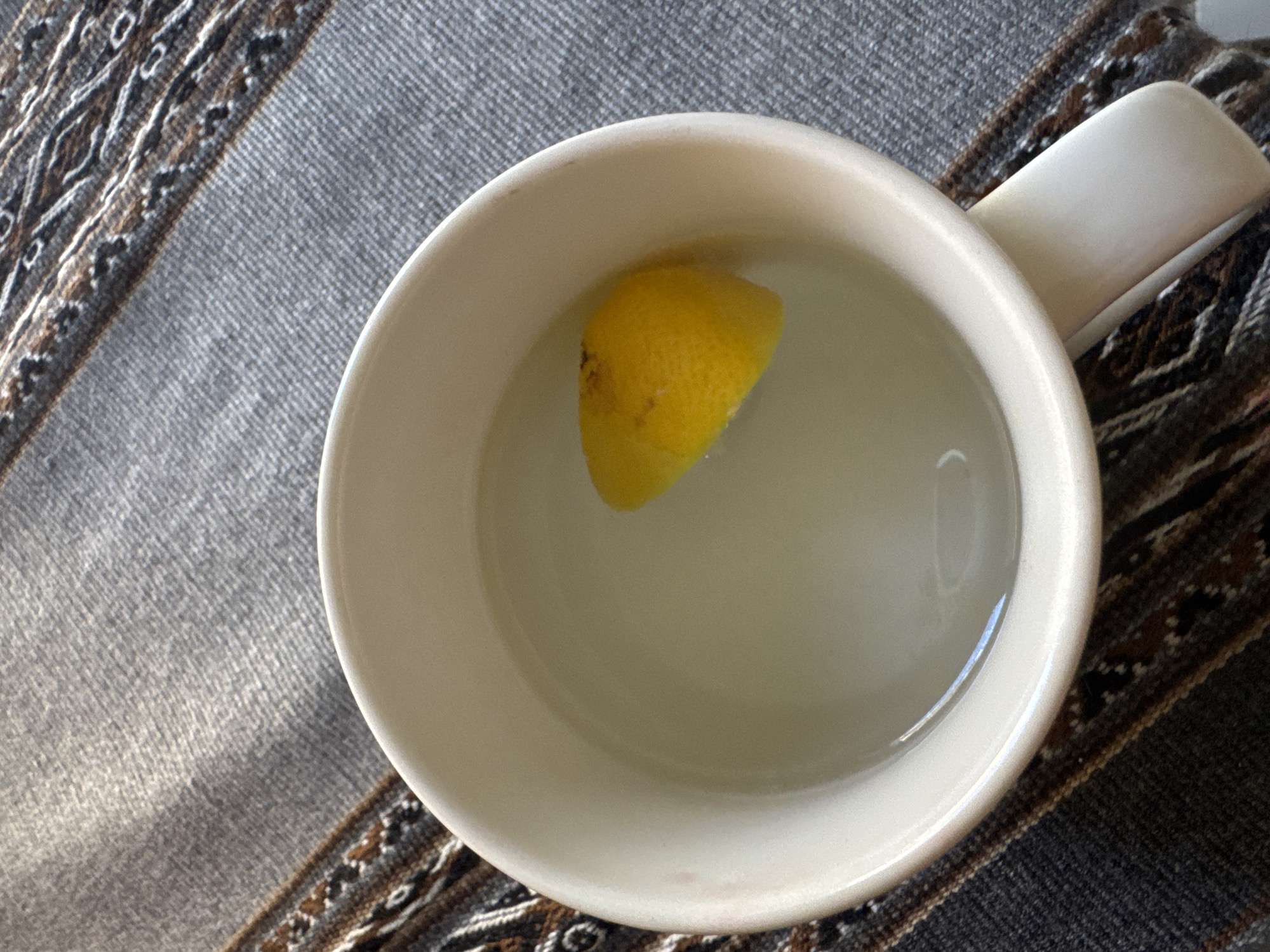 The image captures an overhead view of a white ceramic mug, partially filled with a clear, transparent liquid. A single yellow lemon wedge floats in the liquid, positioned slightly off-center towards the right. The mug has a visible handle extending from its right side. The mug rests on a woven textile surface, likely a placemat or tablecloth. This textile features a predominantly gray, tightly woven pattern, interspersed with a decorative border composed of brown, black, and white geometric designs, running diagonally across the scene from the upper right to the lower left. The lighting suggests ambient illumination from an unseen source, casting subtle shadows and highlights. The scene is located in the city of Unknown, Unknown.