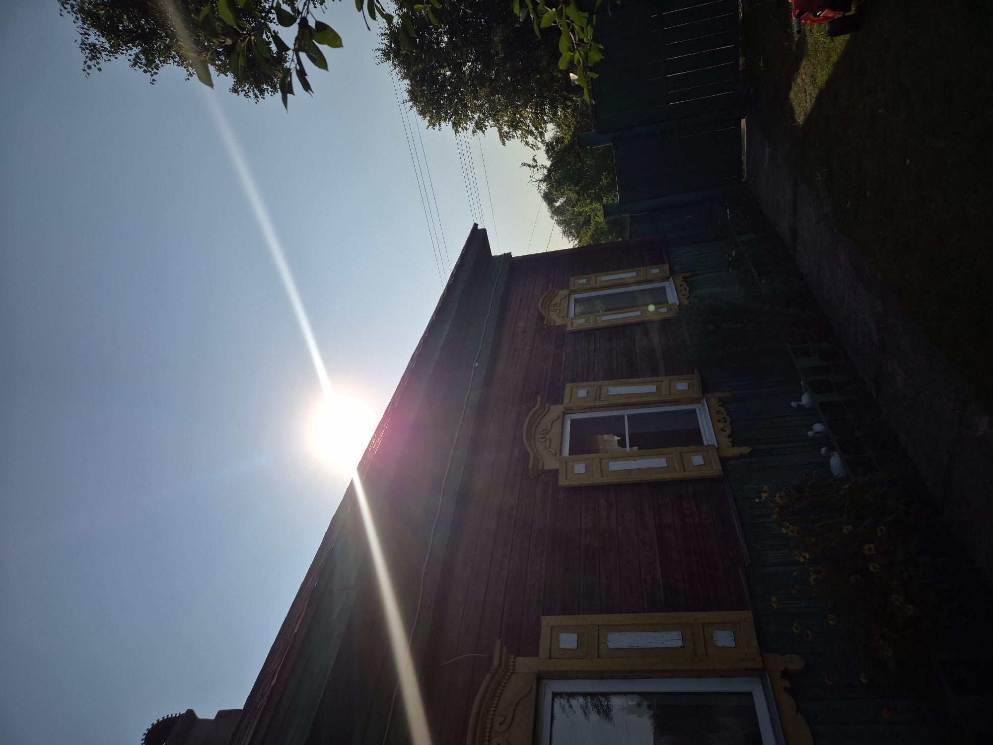 A low-angle, full outdoor shot captures the side of a rustic, wooden house with an angled roof in the village of Parytski Sel'ski Save, Belarus. The sun, positioned high and to the left of frame, is intensely bright, creating strong lens flare that streaks diagonally across the sky and down the side of the house. The house itself is constructed of horizontal dark brown wooden planks, with a dark green painted upper trim and a dark green lower foundation. Two windows are visible on the side of the house, each framed by ornate yellow trim and featuring yellow shutters with white vertical slats. Below the windows, a band of dark green painted wood lines the base of the house, partially obscured by foliage. In the foreground, to the right, a dark green wooden fence and gate are visible, casting long shadows across a grassy yard. To the far right of the frame, a small portion of a red object, possibly a toy or piece of equipment, is visible on the grass. Trees with lush green foliage are present in the background, with visible power lines stretching horizontally across the sky behind the house. The scene is illuminated by the bright sunlight, casting the lower portion of the yard and the side of the fence into shadow.