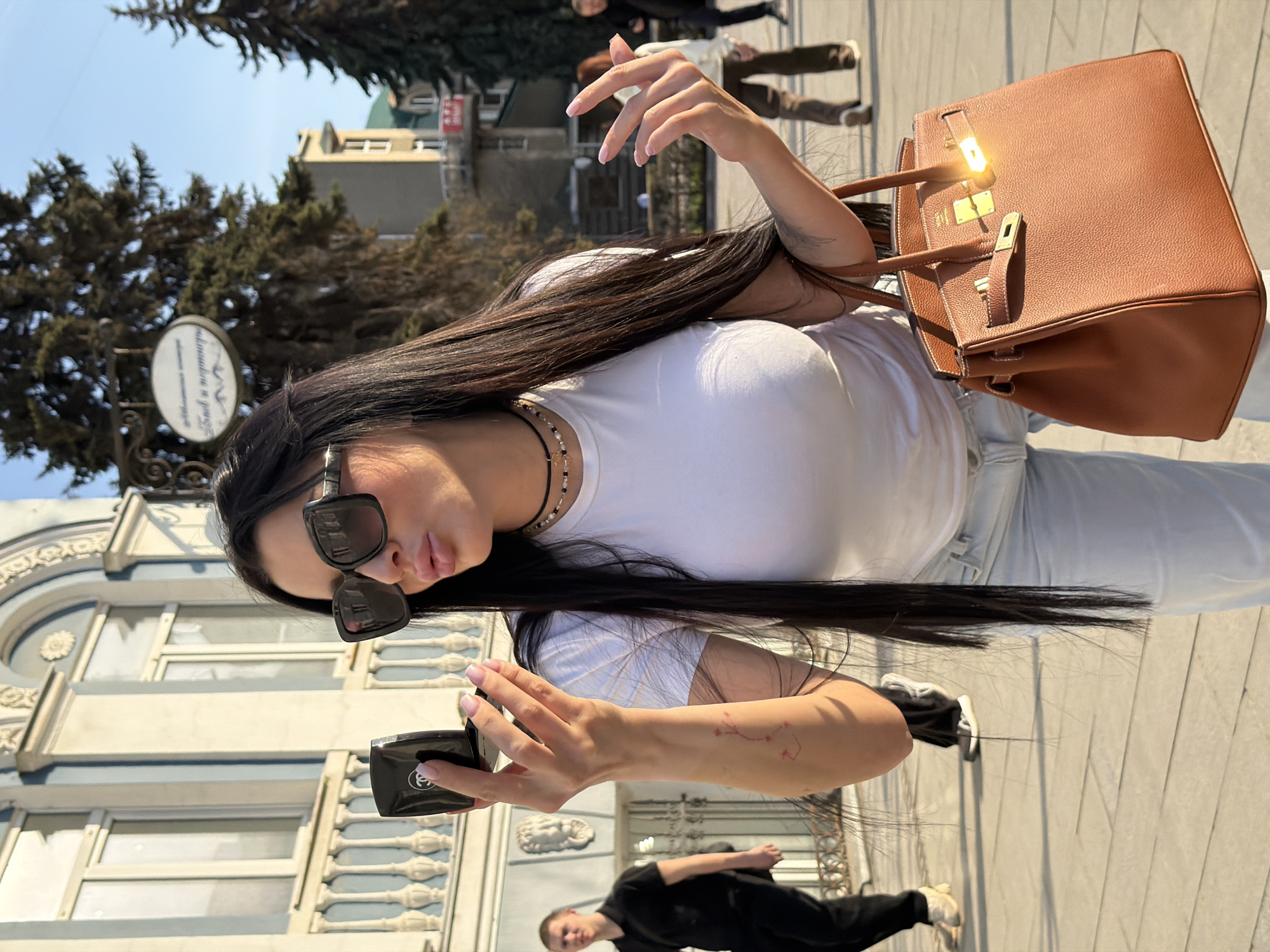 A woman stands outside a building in Voronezh, Russia, wearing sunglasses, a white t-shirt, and light-colored jeans. She holds a compact in her left hand. A brown leather purse hangs on her right shoulder. A man in dark clothing walks near her. Trees and buildings are visible in the background.