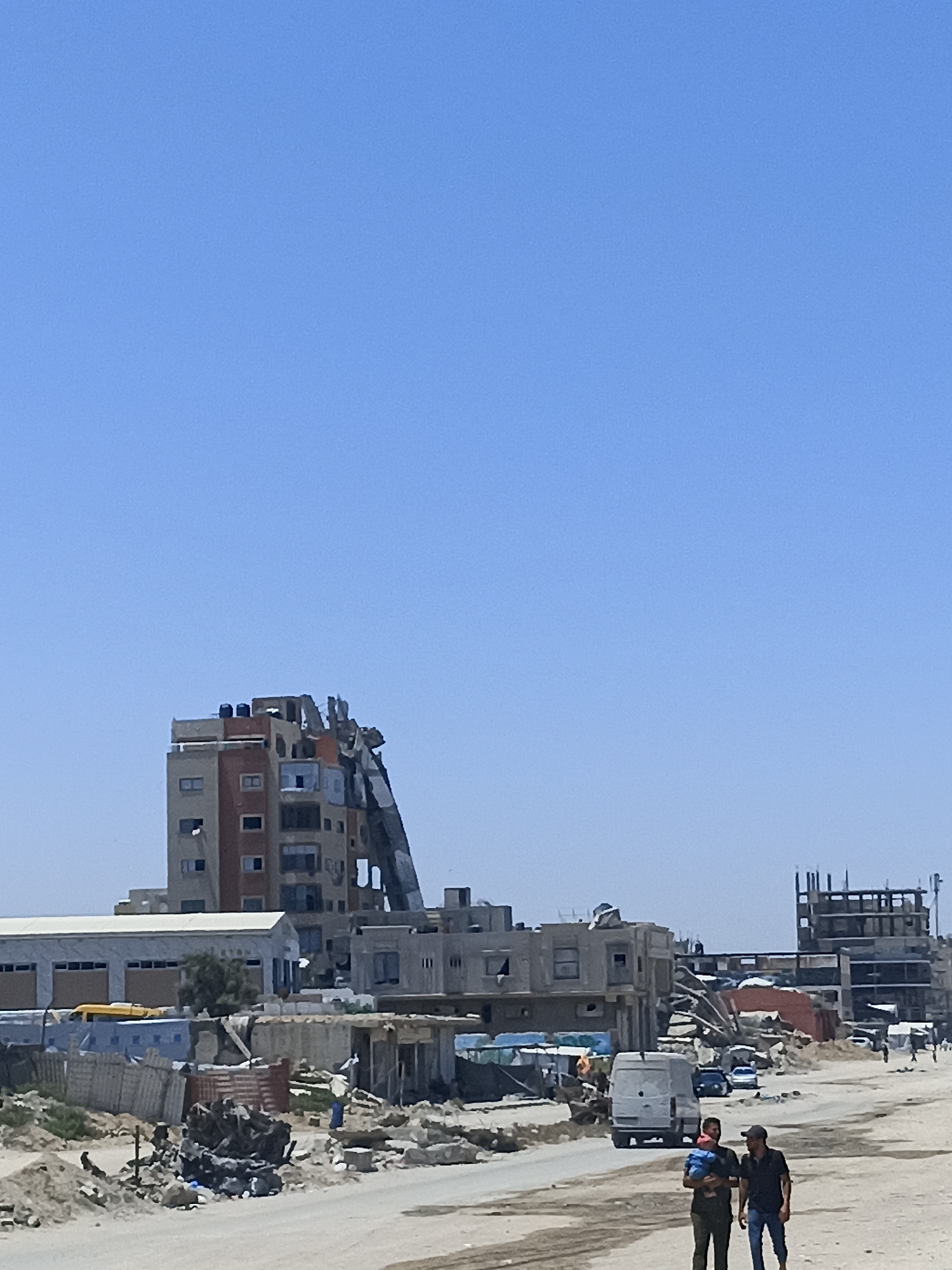 An aerial view of damaged buildings in Gaza City, Gaza Strip, Palestine, depicts a scene of urban devastation under a clear blue sky. In the foreground, a dirt road is visible, with debris scattered along its edges, suggesting recent destruction. Two men are walking on the road, one carrying a baby. Behind them, a white van is parked. Further down the road, several cars are seen.
The middle ground is dominated by a severely damaged multi-story building, its facade partially collapsed, revealing exposed interior structures. Adjacent to it are other buildings, some appearing intact and others showing signs of damage. A long, tilted structure, possibly a ramp or a section of a collapsed building, extends from the damaged building downwards. In the distance, a partially constructed building with visible scaffolding stands as a stark reminder of ongoing development amidst the destruction. The overall environment appears arid and dusty, consistent with the Gaza Strip's landscape.