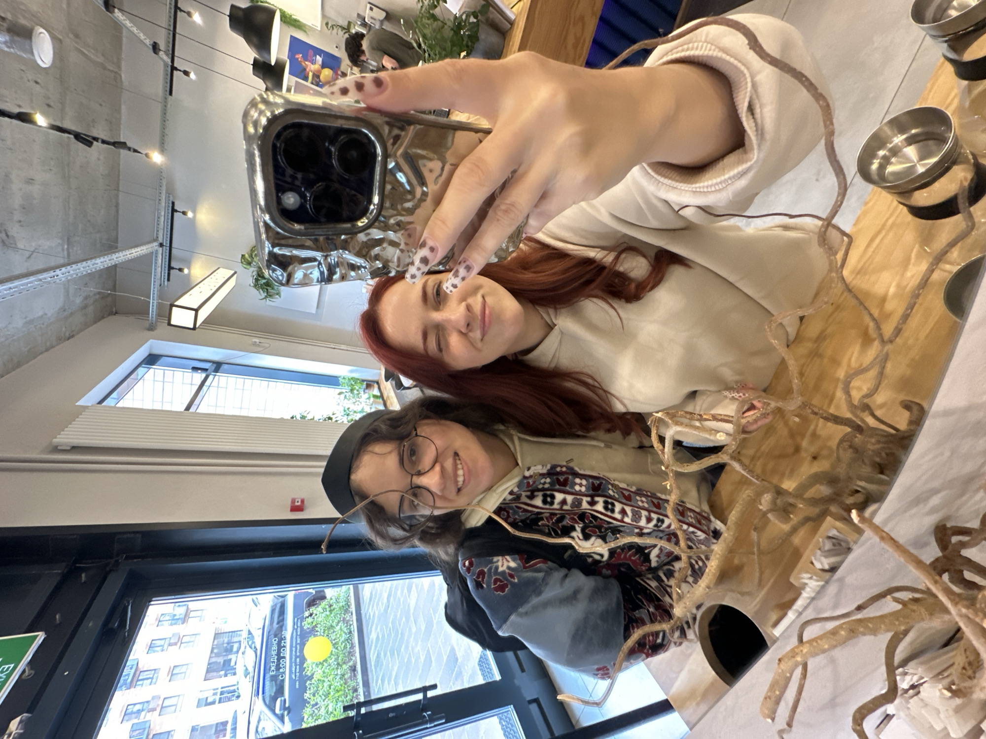 Two young women are taking a selfie in what appears to be a cafe or restaurant. The woman holding the phone has red hair and is wearing a light-colored sweatshirt. The other woman has dark hair, is wearing glasses and a black cap, and is dressed in a patterned jacket. The background shows a window with a view of buildings and greenery outside. A wooden table is visible with decorative objects and metal containers.