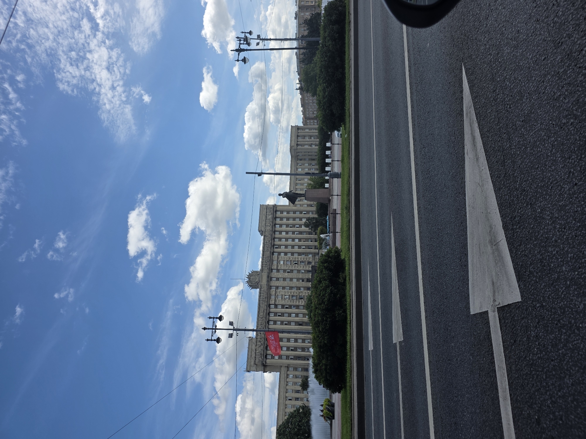 The image shows a wide asphalt road with white directional arrows painted on it. In the background, there is a large, imposing building with classical architectural elements, including columns and pediments. A statue of a man in a coat stands in front of the building. A red flag with Cyrillic lettering is visible on a flagpole. The sky is blue with scattered white clouds. The scene appears to be a public square or avenue in Saint Petersburg, Russia, likely a significant historical or governmental area given the monumental architecture and statue. There are no visible people in the image, and the action depicted is static, focusing on the architecture and urban landscape.