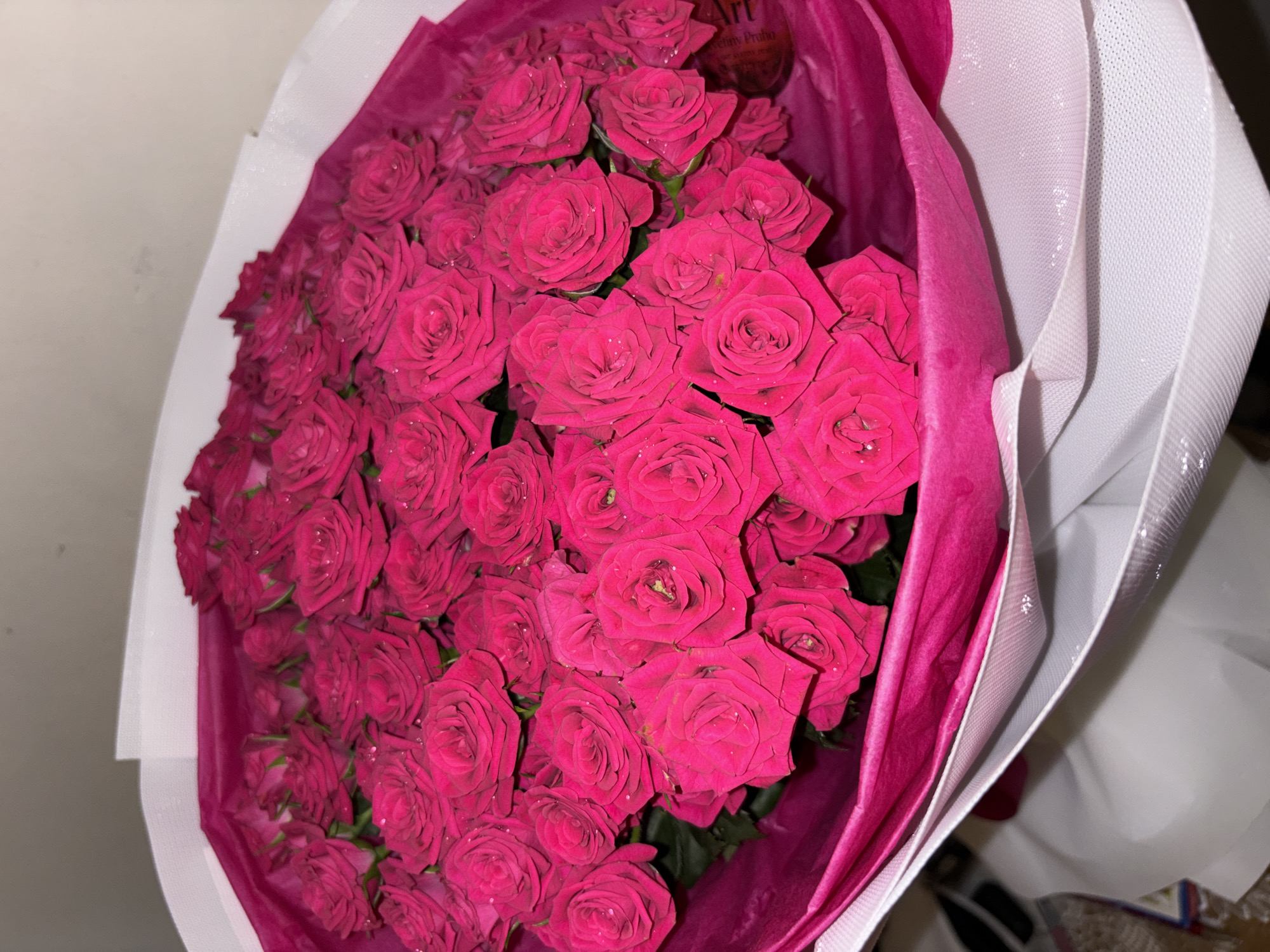 A close-up, high-angle shot shows a large bouquet of vibrant pink roses, tightly packed and wrapped in layers of pink and white paper. The roses, some with visible water droplets, are the primary subject, filling the frame. The background is a plain, neutral color, with the bottom portion of the image showing the white paper wrapping extending downwards. There is no indication of a location in Prague, Czechia, or any specific environmental details. The image is static and focuses solely on the floral arrangement.