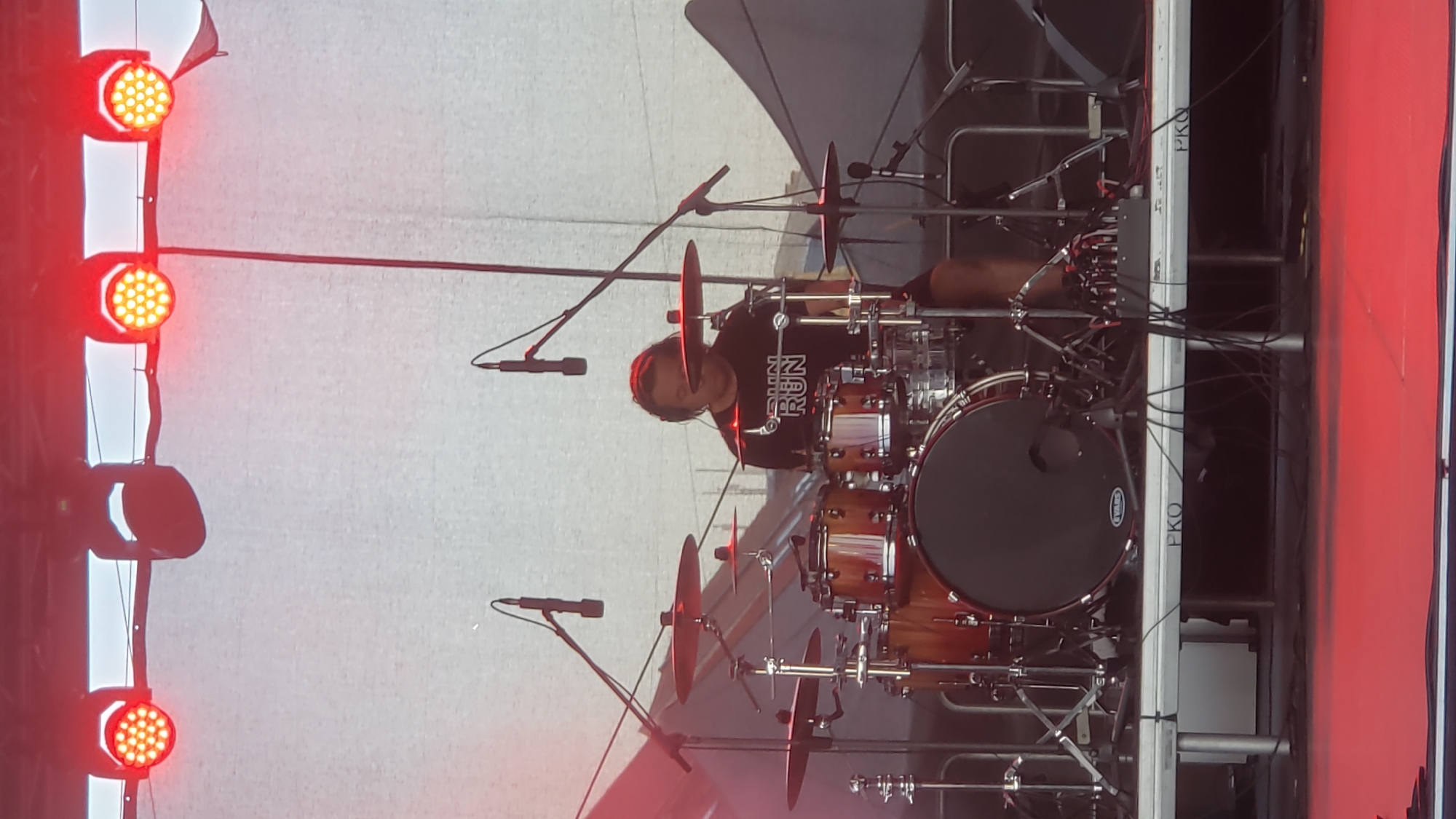 The image depicts a drummer playing a drum set on a stage. The drummer is wearing a black t-shirt with white lettering that reads "RUN RUN". The drum set is composed of several drums and cymbals. Two microphones are positioned above the drum set. Overhead, there are several red stage lights illuminating the scene. The background consists of a light-colored curtain and some darker fabric elements, possibly stage rigging or tents. In the foreground, the edge of the stage is visible with the text "PKO" printed on it, suggesting the location is the PKO Amphitheater in Prešov, Slovakia. The stage is lit with red lighting at the bottom, creating a dynamic atmosphere. The drummer appears focused on playing.