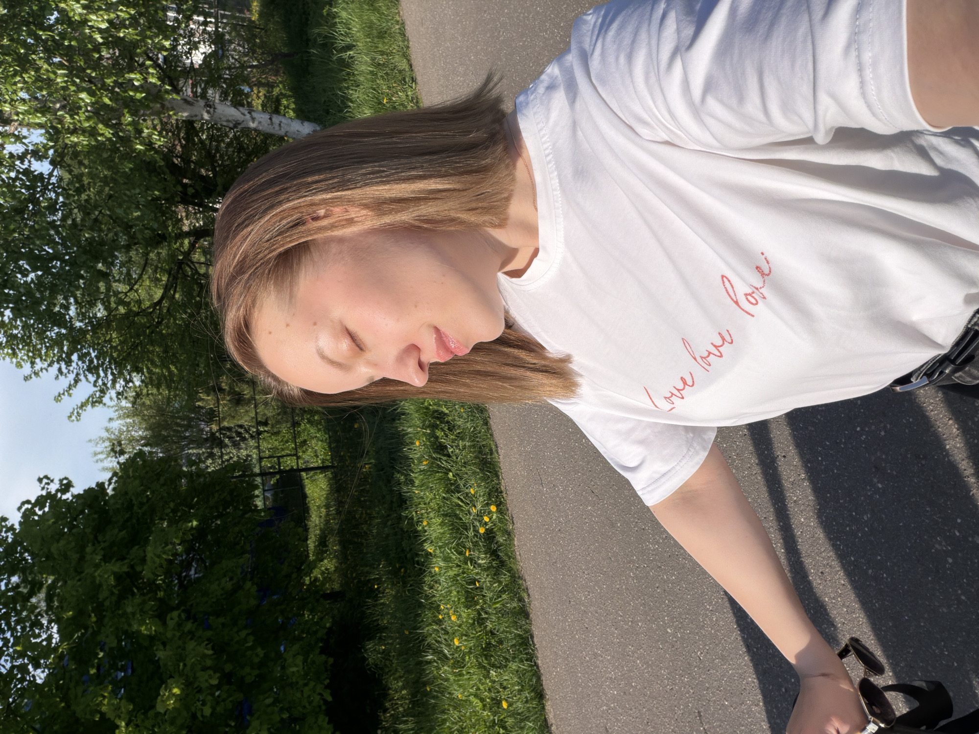 A woman with light brown hair is outdoors on a sunny day in what appears to be Reshetnikovo, Russia. She is wearing a white T-shirt with red cursive writing, partially obscured, possibly saying "love love love". She is standing on a paved surface next to a grassy area with dandelions. A tree with white bark is visible in the background. The image is taken at an angle, suggesting she might be holding the camera herself, and has her eyes closed. She is holding sunglasses in her hand.