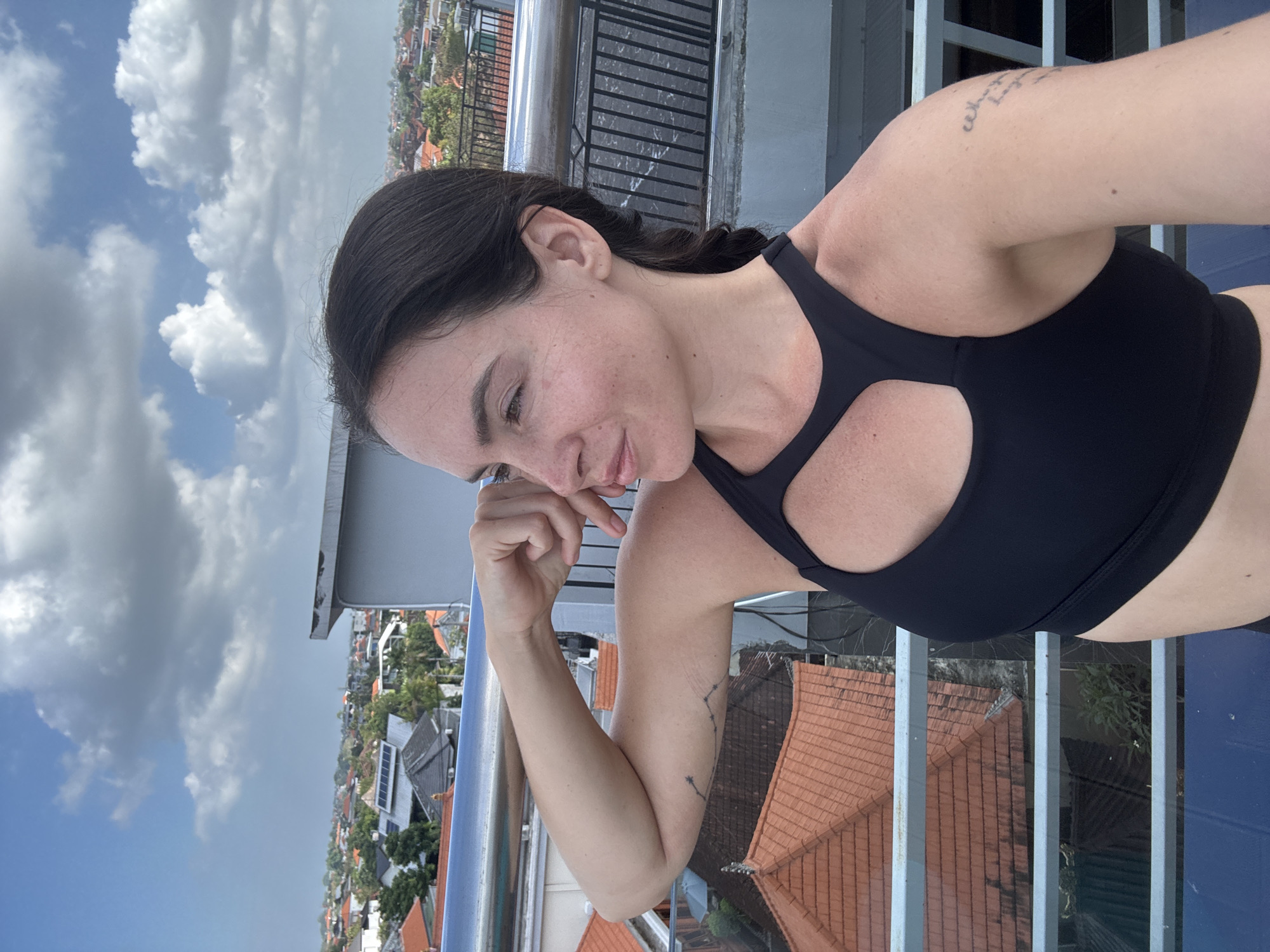 In North Kuta, Indonesia, a woman with dark hair pulled back in a braid stands on a rooftop, her left arm flexed with her hand near her face. She wears a black sports bra. The background shows a cityscape with red-tiled roofs and a cloudy sky. A metal railing is visible in the foreground.