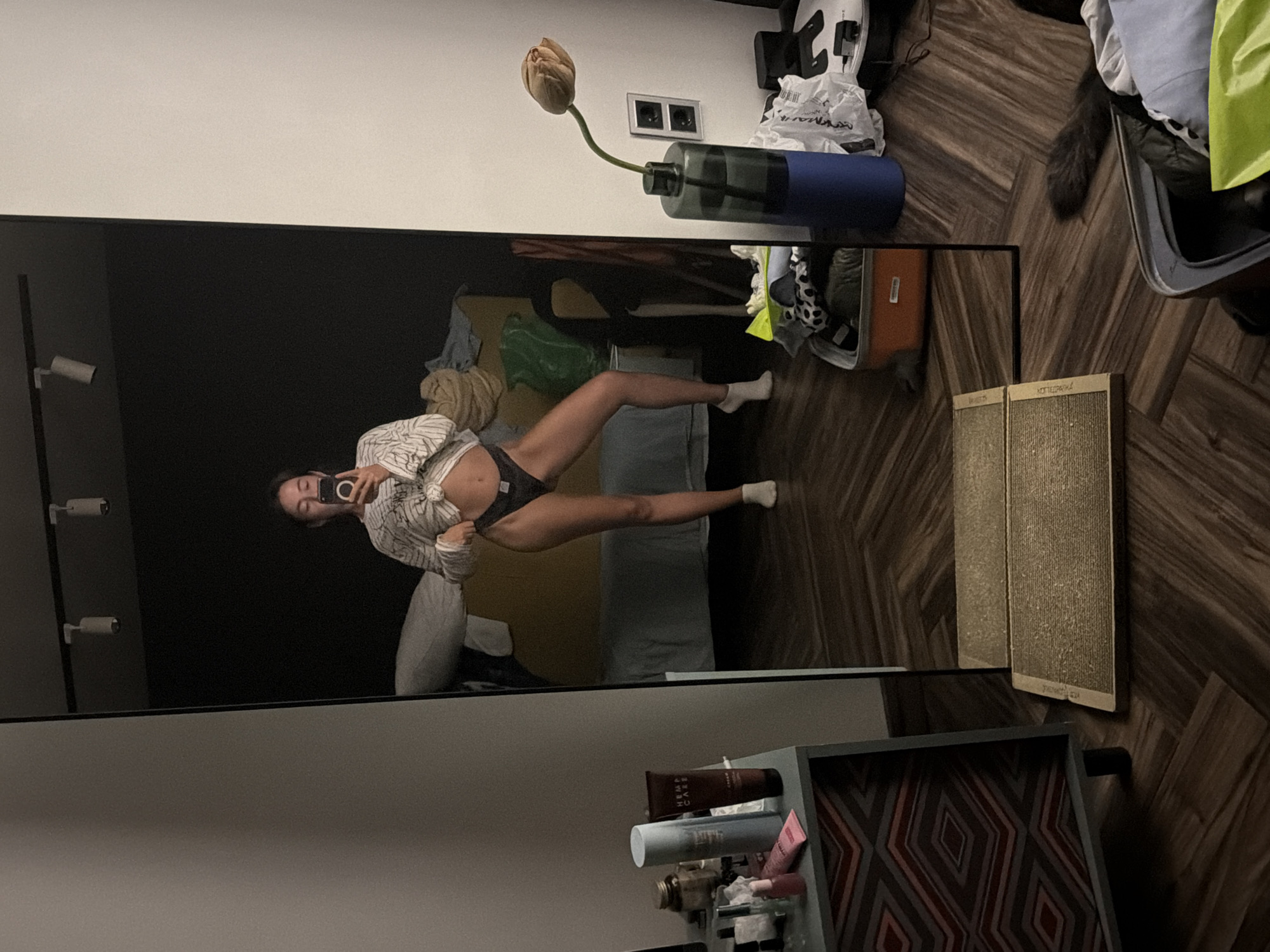 The image shows a young woman taking a mirror selfie in what appears to be a bedroom. She is wearing a white striped shirt, partially untied to reveal her midriff, and black underwear. She is standing in front of a large, full-length mirror. The room is somewhat cluttered. To the left of the mirror is a dresser with various cosmetic items on top. In the background, visible in the mirror's reflection, is a bed with a yellow headboard and a dark wall. To the right of the mirror, there is a vase with a single dried flower, a dark blue cylindrical object, some plastic bags, and a suitcase. The floor is made of dark wooden planks laid in a herringbone pattern. A portion of what appears to be a cat's tail is visible on the floor to the right of the mirror. The setting does not provide any specific visual cues to place it in Ust-Ilimsk, Russia, or any other particular city.