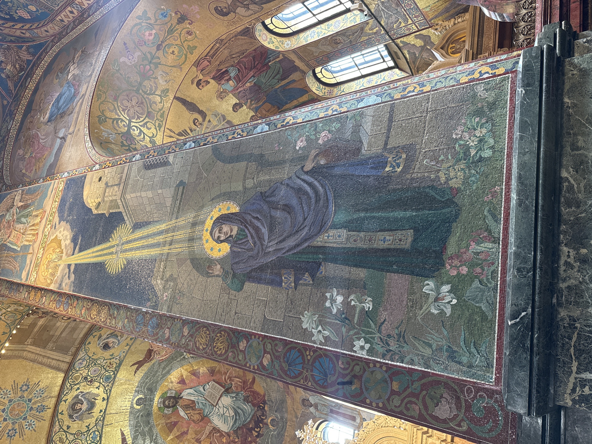 The image shows an interior view of the Church of the Savior on Spilled Blood, located in Saint Petersburg, Russia. Visible are ornate mosaic panels and ceilings typical of Russian Orthodox church design. The primary mosaic in the image depicts a religious figure, possibly the Virgin Mary, with a halo and surrounded by light rays and a floral scene. Other mosaic panels feature additional figures and decorative patterns. There are visible architectural details, including windows and marble features.
