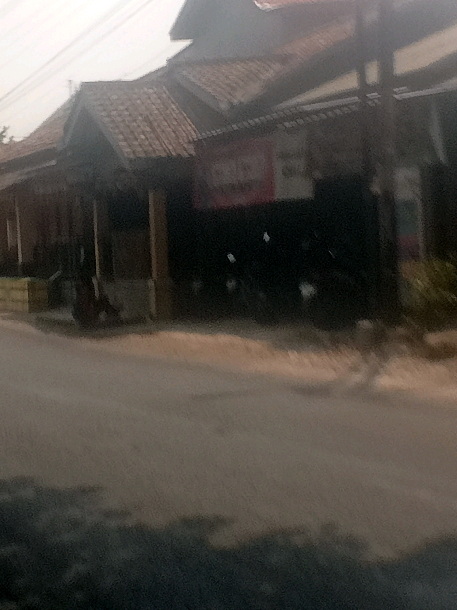 The image depicts a slightly blurred outdoor scene with a building that appears to be a shop or restaurant in Lebakgowah, Indonesia. The building has a tiled roof and a covered outdoor area supported by wooden posts. In front of the building, there is a street and what appears to be a wall or low barrier made of yellow bricks. Several motorcycles are parked under the covered area. The overall lighting suggests it is daytime, possibly late afternoon, with soft, diffused light. The environment appears to be a relatively quiet street with minimal activity visible.