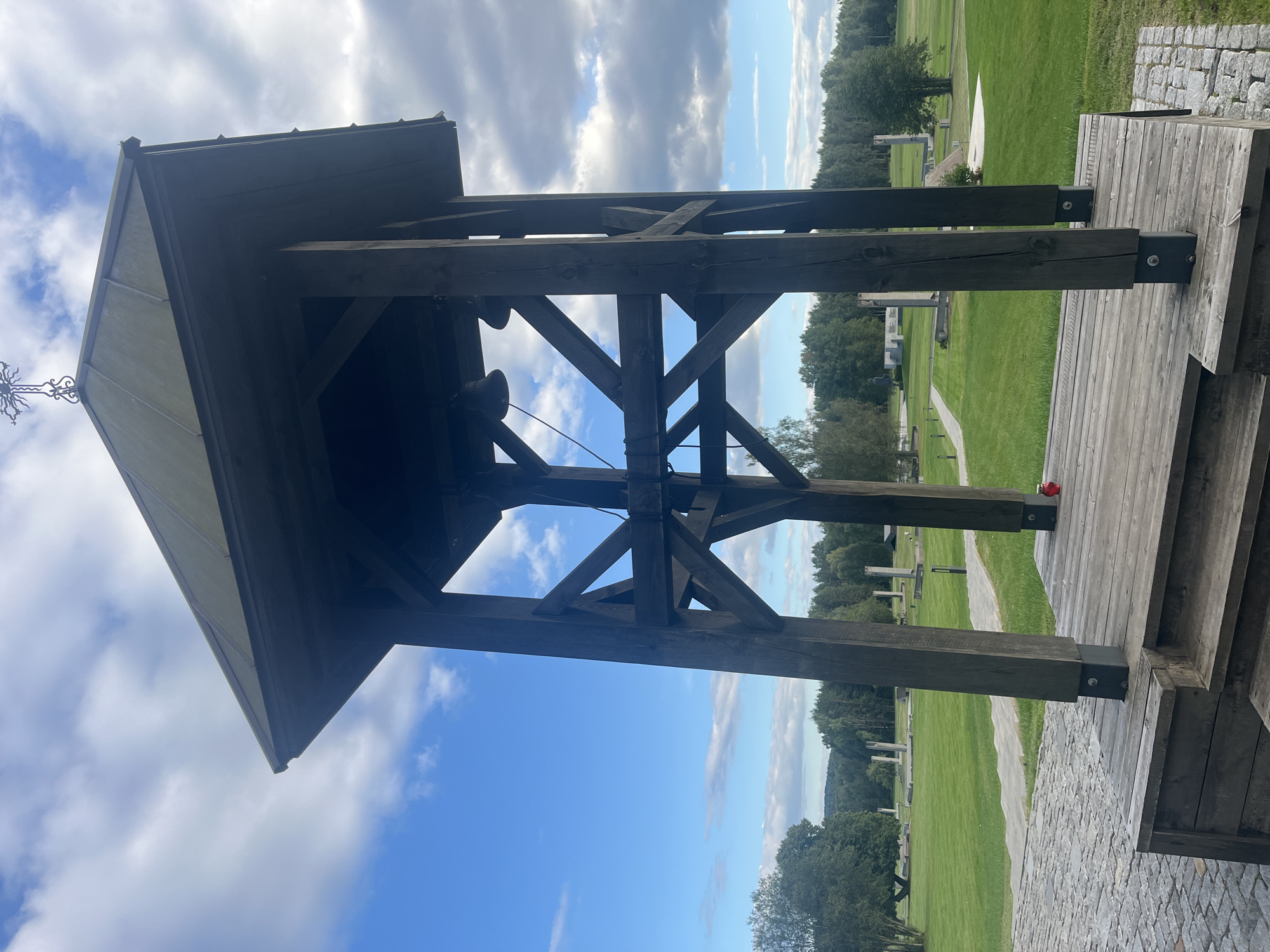 This is a low-angle, medium shot of a wooden bell tower with a cross on top, set in a grassy field under a partially cloudy sky. The bell tower is constructed of dark, weathered wood and has a sloped, dark roof. Two bells are visible hanging within the structure. The base of the tower is a wooden platform with steps. In the foreground, to the left, is a paved area made of cobblestones. In the middle ground and background, the field is dotted with rows of white, pillar-like structures, some of which are accompanied by plaques or inscriptions, suggesting a memorial site. Trees form a dense line along the horizon. A single red memorial candle sits on the wooden platform in front of the bell tower. The location is identified as Kamyanyetsky Selsoviet, Belarus.
