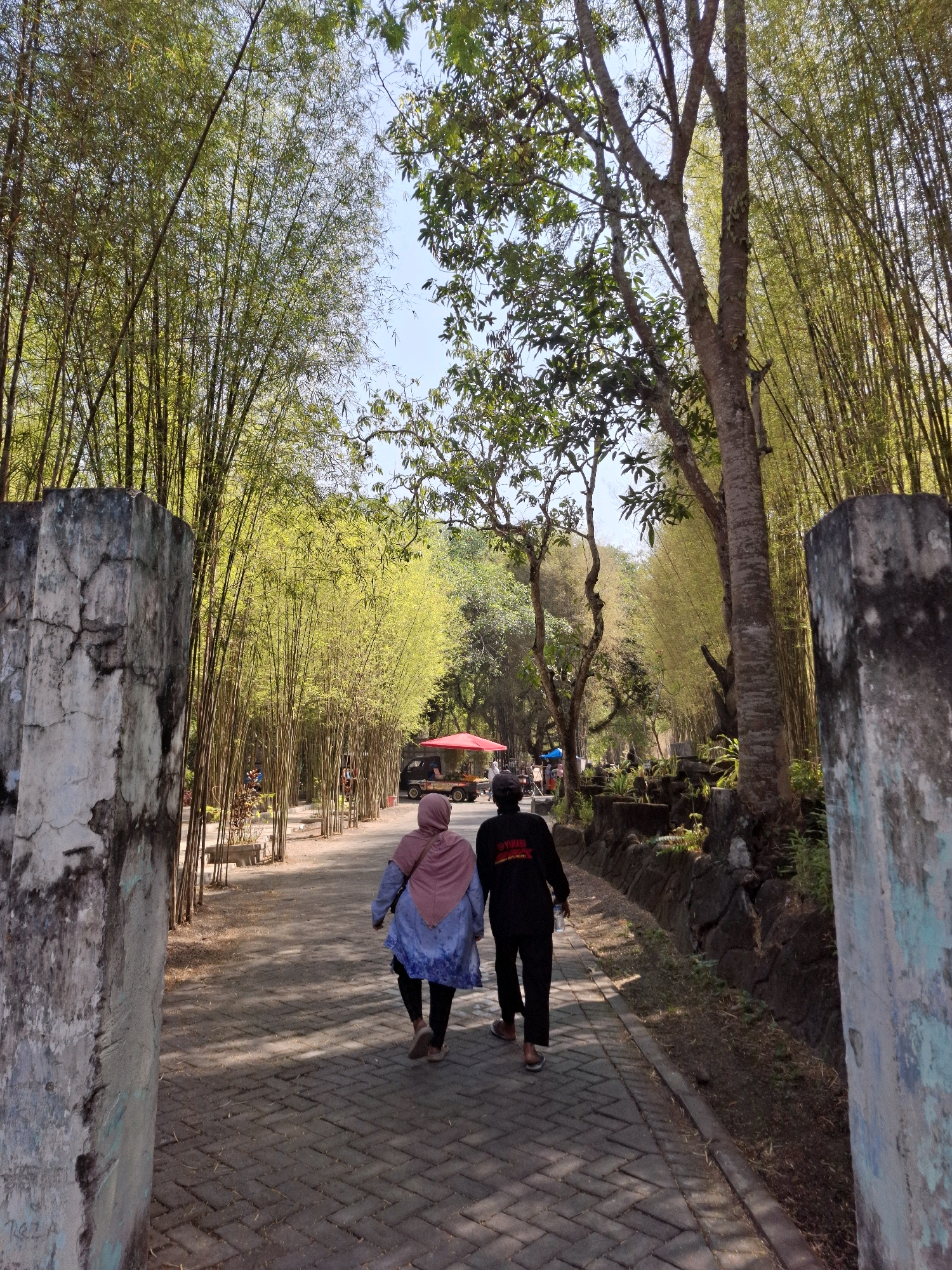 The image shows a pathway lined with bamboo trees in Tawun, Indonesia. Two individuals, a woman wearing a pink hijab and a blue and white patterned tunic, and a man in a black jacket with a Yamaha logo, are walking away from the camera down the path. The path is paved with interlocking bricks and is bordered by stone walls and vegetation. In the background, there is a red umbrella covering a vendor's stall and a vehicle. The sky is clear and blue, and the sunlight casts shadows on the path. The overall scene depicts a serene, outdoor environment.