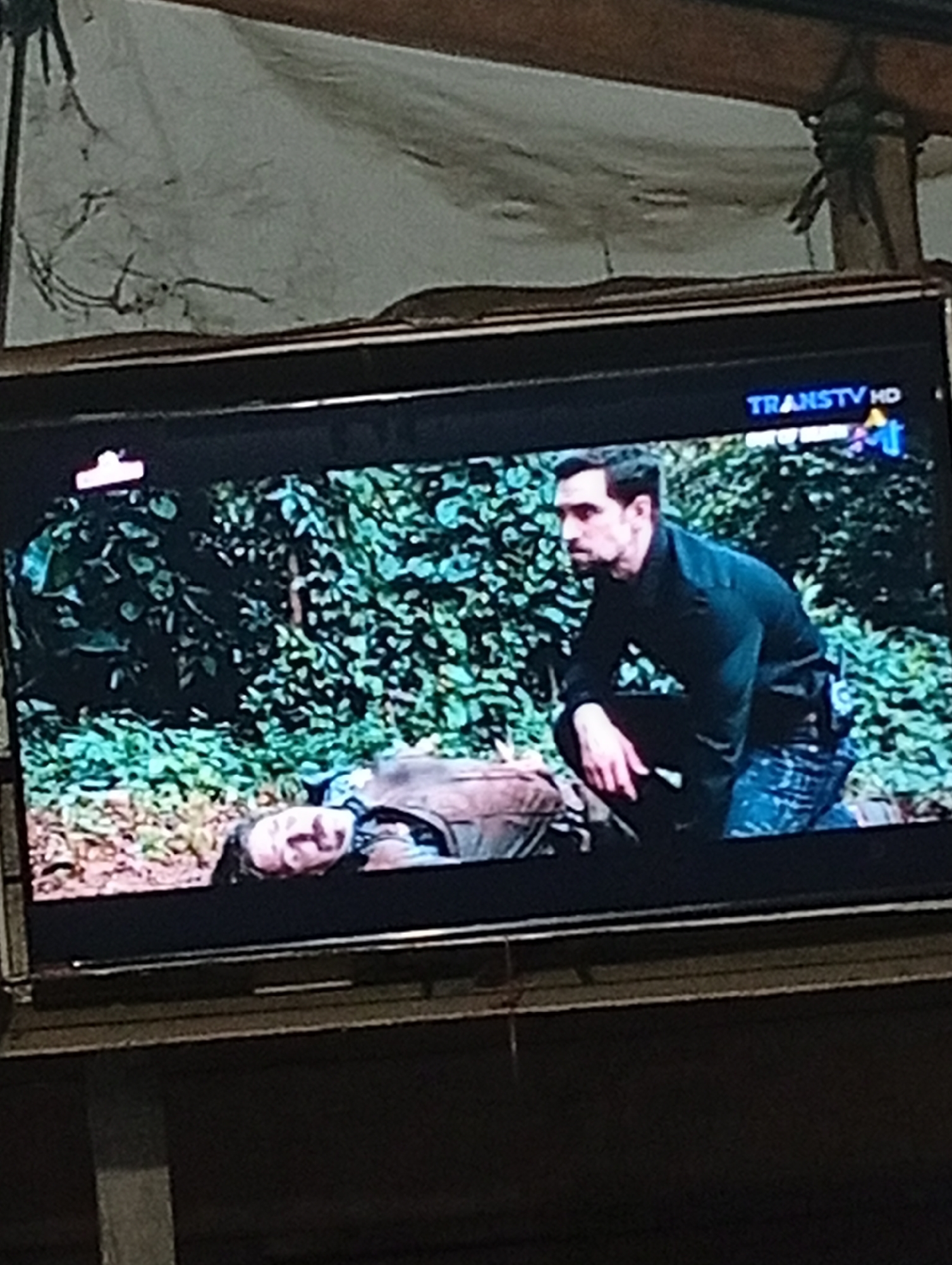The image shows a television screen displaying a scene from a television program. The program appears to be a drama or action series, with two men featured. In the foreground, a man with dark hair and a beard is crouched down, looking to his left with a serious expression. He is wearing a black long-sleeved shirt and blue and white patterned pants. Behind him and to his left, another man is lying on the ground, appearing to be injured or unconscious. He is wearing a brown jacket over a lighter colored shirt. The background is filled with lush green foliage, suggesting an outdoor, possibly jungle or forest setting. The TRANS7 HD logo is visible in the upper right corner of the television screen. The television itself is mounted on a wall or stand, and the surrounding environment is dimly lit, with a textured, possibly patterned ceiling visible above the screen. The image does not contain any specific visual information that would allow for the determination of the city of Lhokseumawe, Indonesia.
