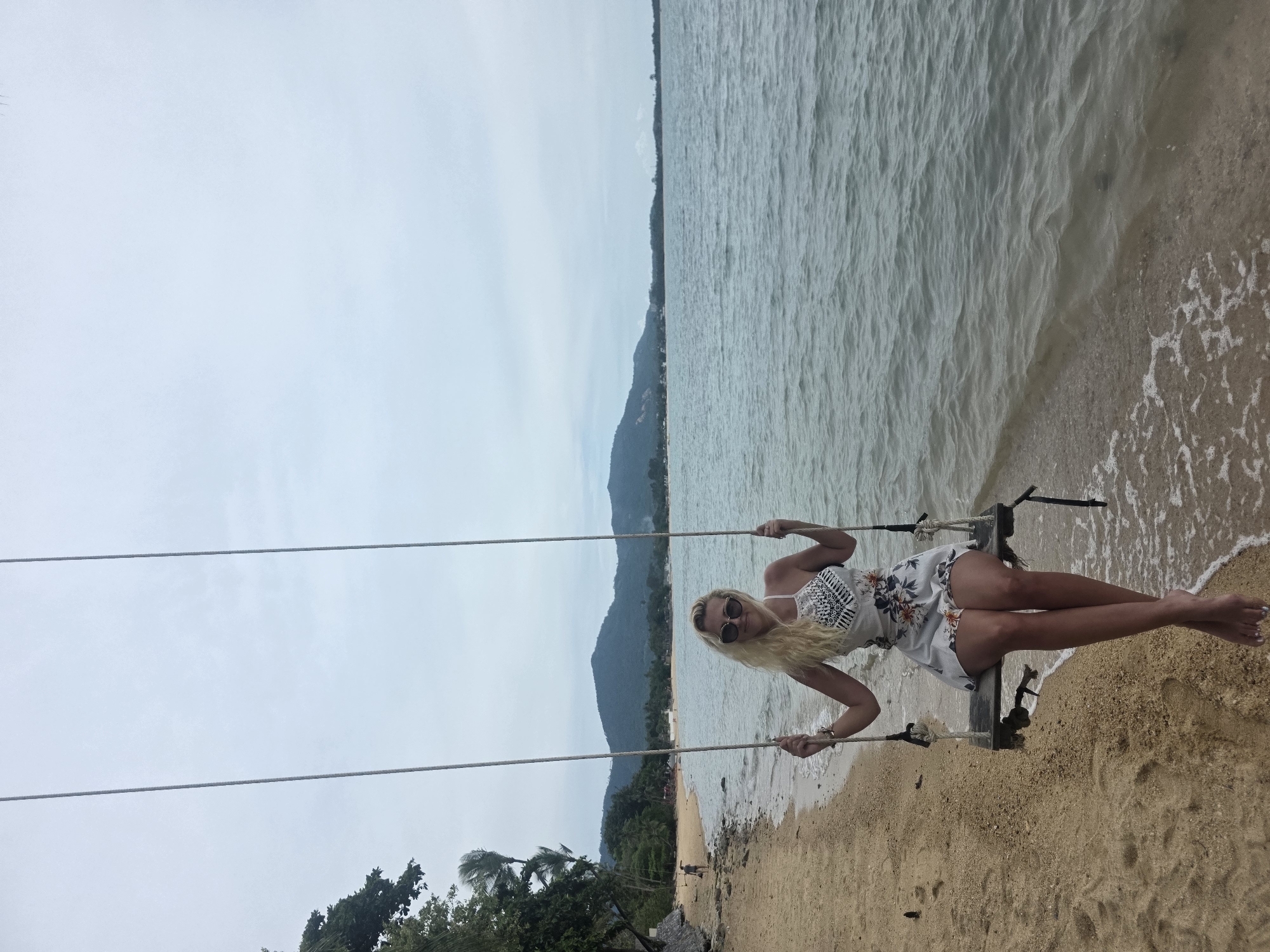 A blonde woman is sitting on a swing on a beach in Baan Bo Phut, Thailand. The swing is made of wood and rope, and it is suspended from two ropes. The woman is wearing a white floral dress and sunglasses. She is smiling and looking at the camera. The beach is sandy and there are palm trees in the background. The ocean is calm and there are mountains in the distance. The sky is cloudy.
