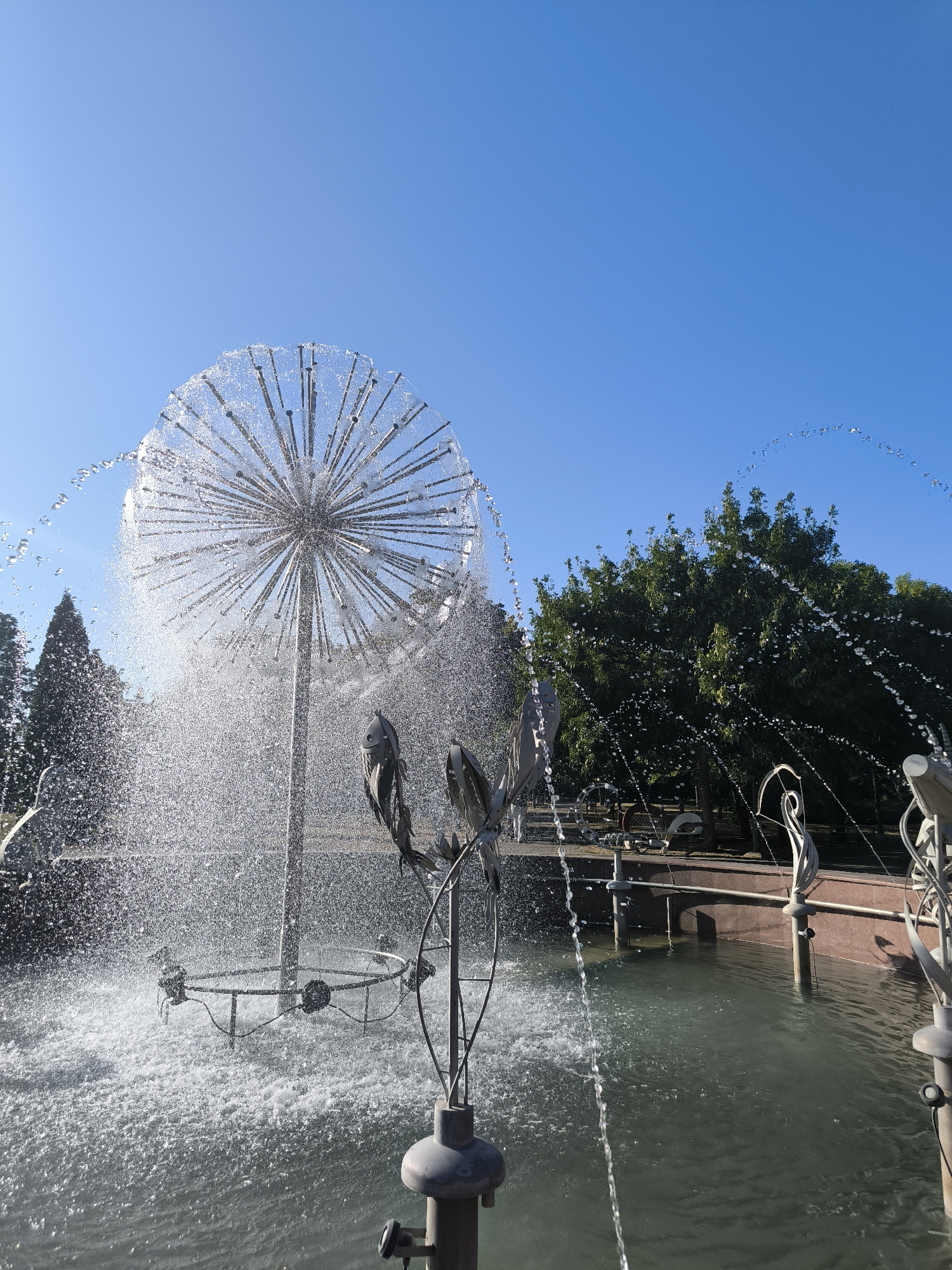 The image depicts a modern fountain with a spherical metal structure that sprays water in a dandelion-like pattern against a clear blue sky. The fountain is situated in a pool of water, and decorative metal sculptures are visible in the foreground and around the pool's edge. Lush green trees form the background, with a hint of a park-like setting. The scene appears to be a public space, possibly a park or plaza. This fountain is located in Chișinău, Moldova, specifically in the Valea Morilor Park. There are no people visible in the image.