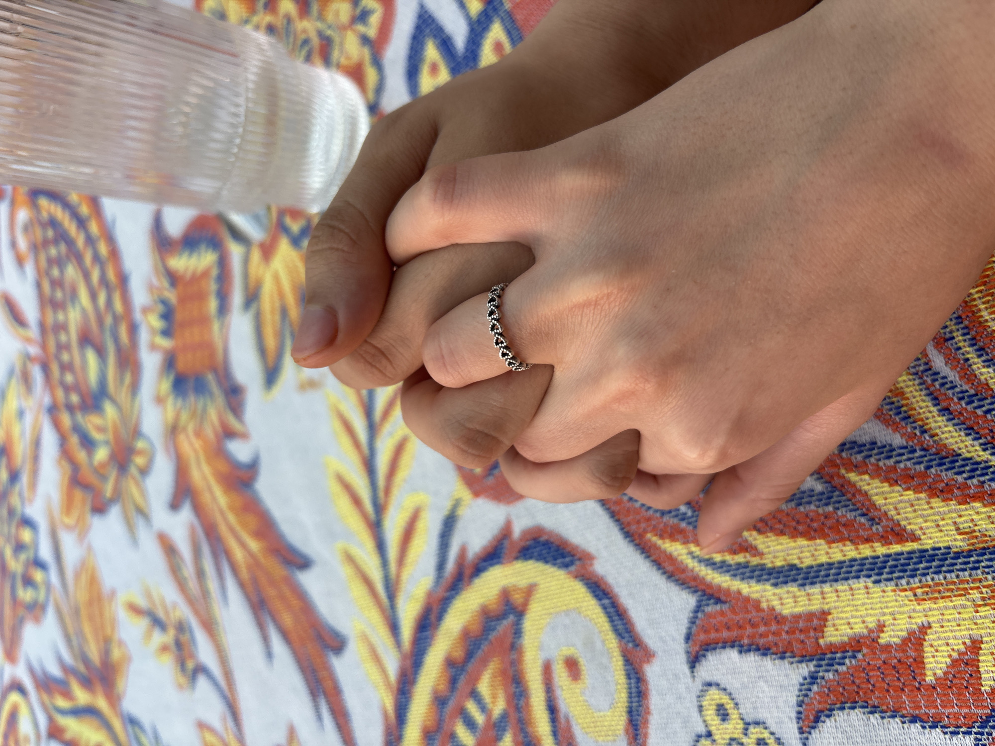 The image shows two hands intertwined, resting on a patterned tablecloth. The tablecloth displays a repeating design of yellow, blue, orange, and red paisley-like elements against a white background. A transparent, ribbed glass is partially visible in the upper left corner. The hand on the right wears a ring adorned with small heart-shaped details. The scene is likely indoors, due to the controlled lighting. The location is stated to be Puebla, Mexico, though no specific details in the image directly confirm this.