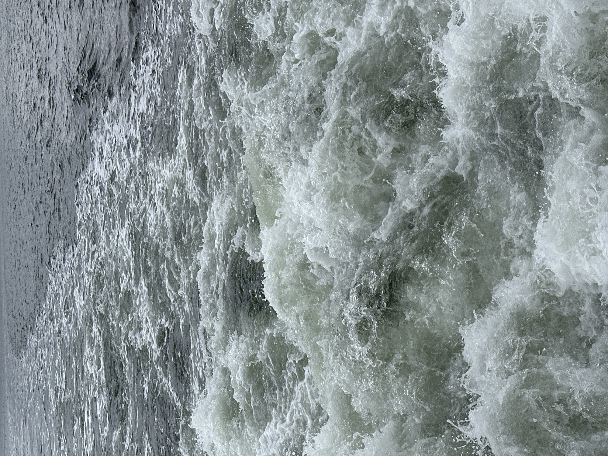 The image captures a turbulent wake of water, likely generated by a moving vessel. The foreground is dominated by frothy white foam and churning waves, suggesting rapid movement. The water transitions to a darker, less agitated surface further in the background. This scene is consistent with the waters of the Kiel Fjord in Kiel, Germany, where the busy maritime traffic often creates such wakes. There are no people or specific objects clearly visible, only the dynamic interaction of water. The environment is solely the aquatic expanse of the fjord.