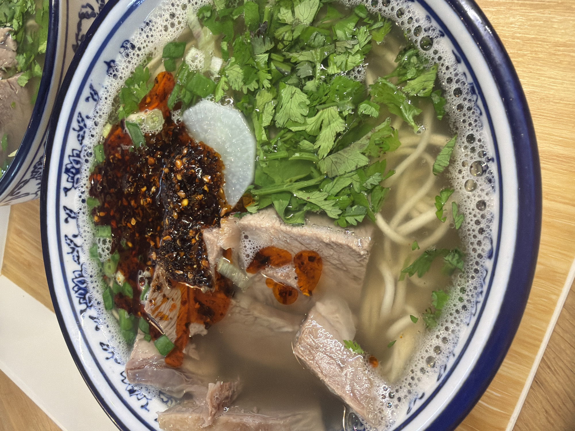 A close-up, overhead shot shows a bowl of steaming beef noodle soup. The soup is clear with noodles, sliced meat, a dollop of chili oil, chopped scallions, cilantro, and a slice of daikon radish. The bowl has a blue and white floral pattern around the rim, and it sits on a light-colored, wooden surface. The image is well-lit and focuses on the appetizing details of the dish. There is no visible context of Saint Petersburg, Russia.