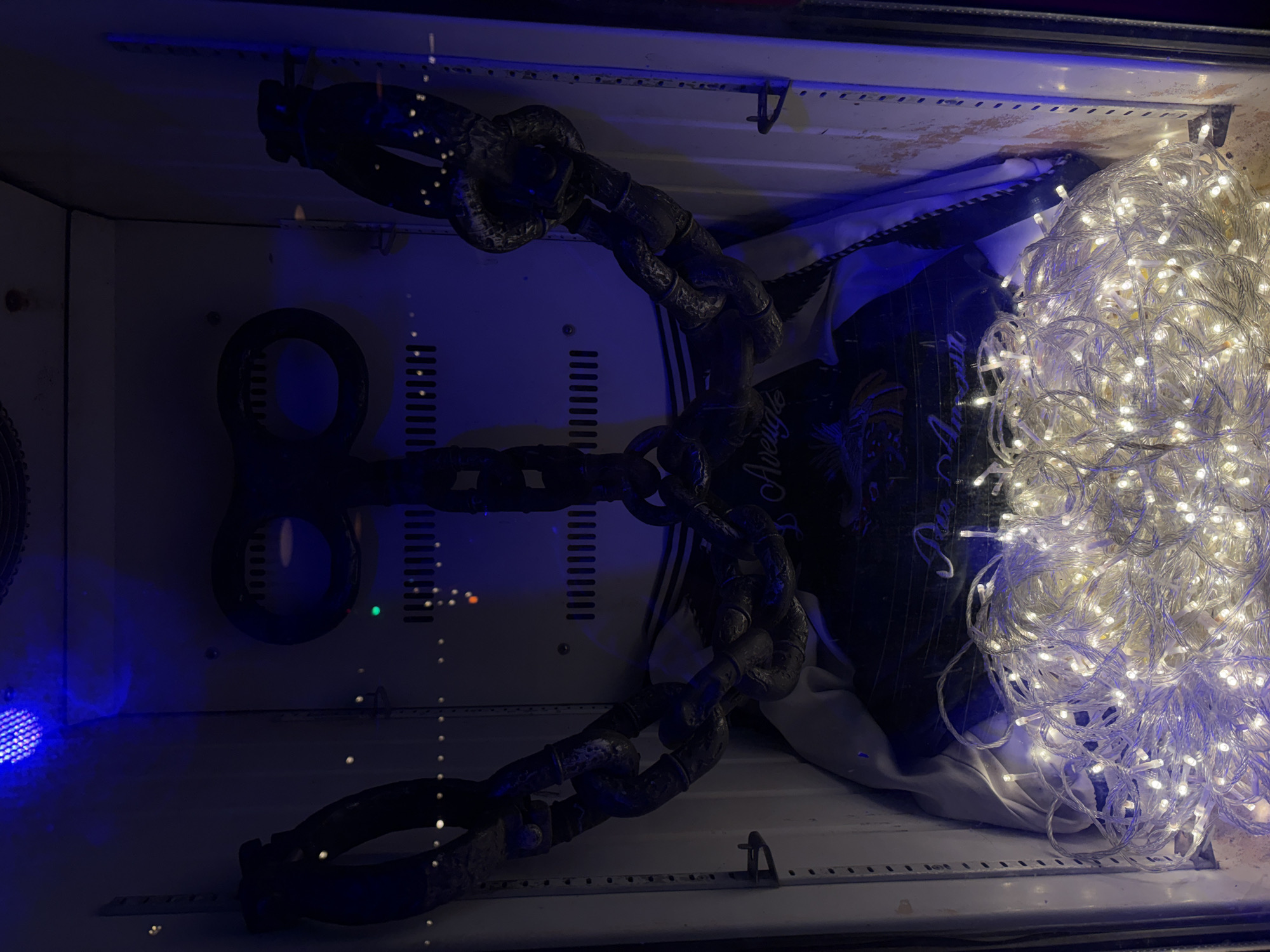 A dimly lit scene, seemingly inside a container in Baan Bang Rak, Thailand, features a large metal chain suspended from the ceiling, obscuring a black garment with partial text. A heap of fairy lights brightly illuminates the right side of the container, while a blue light is visible on the lower left. The container's interior is predominantly white, with visible vents and a metal frame.