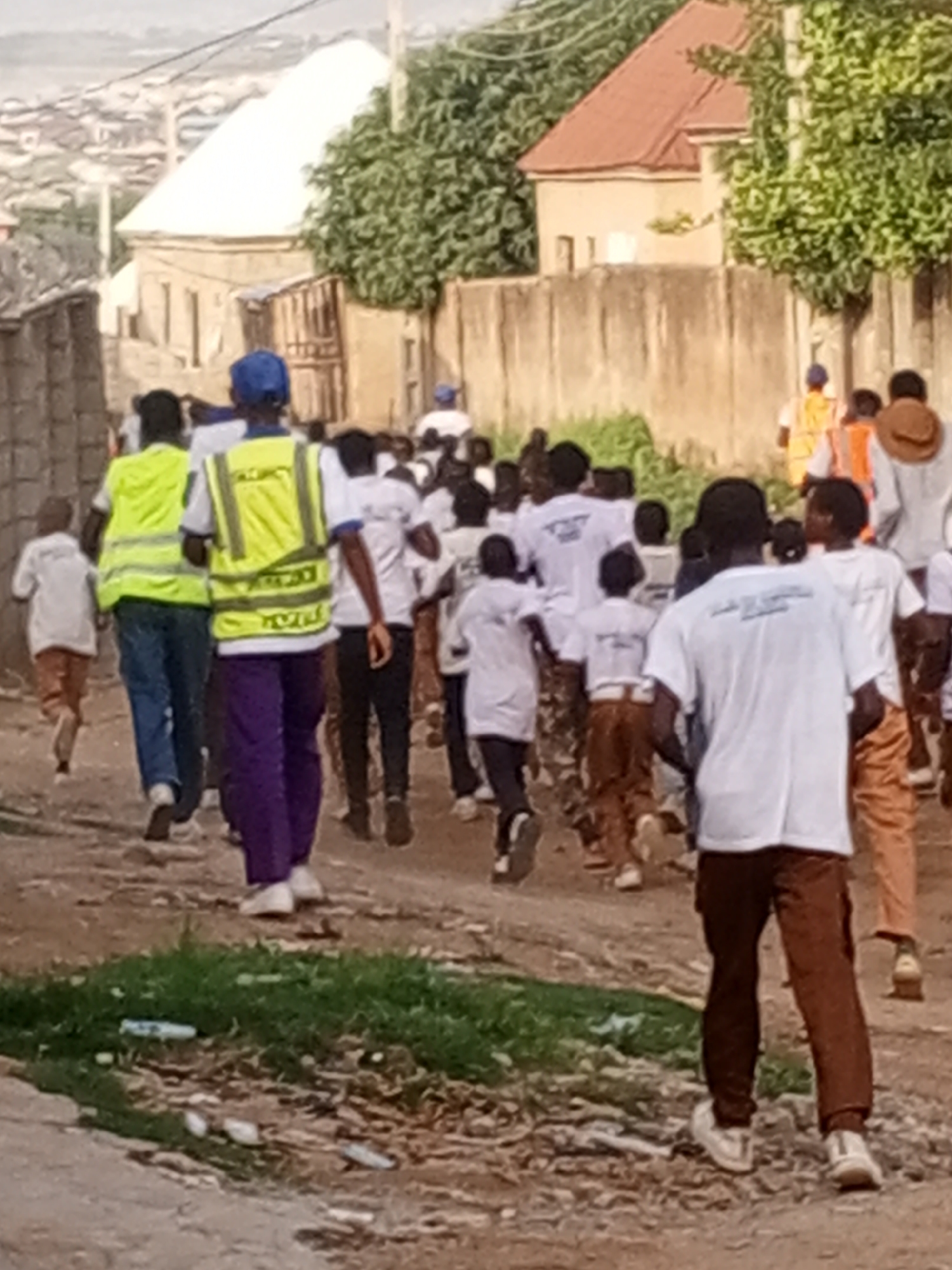 A group of people, possibly school children, are running on a dirt road in a city neighborhood. The people are wearing a combination of white t-shirts, brown pants, and high-visibility vests. Some people are wearing hats. There are residential buildings with walls in the background. The location is the city of Unknown, Unknown.