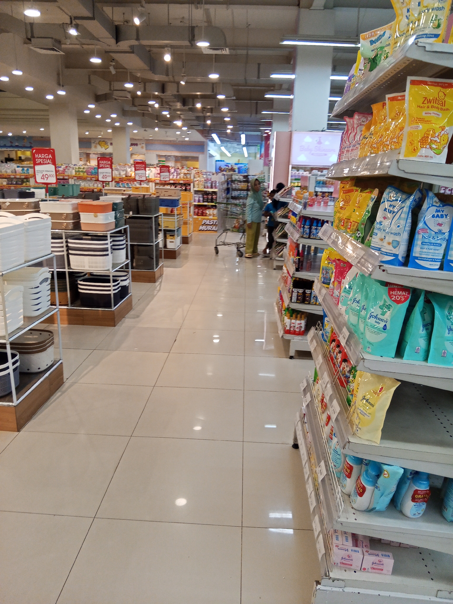 The image shows the interior of a well-lit supermarket in Lubuklinggau, Indonesia. The scene is dominated by aisles of merchandise, primarily household goods and toiletries. In the foreground, shelves are laden with packaged baby products, including shampoo, body wash, and lotions, predominantly from brands like Zwitsal and My Baby. There are also rows of plastic storage containers and baskets on display.
Further into the store, visible in the background, are more shelves stocked with various consumer goods, including food items and cleaning supplies. Several shopping carts are present in the aisles. Two people are visible: a woman wearing a headscarf and a light blue top, pushing a shopping cart, and a child walking behind her. The floor is tiled with a light beige color, reflecting the overhead lighting. Promotional signs, including "HARGA SPESIAL" (Special Price), are displayed on some shelves, indicating ongoing sales. The overall environment suggests a typical retail setting within a city like Lubuklinggau.