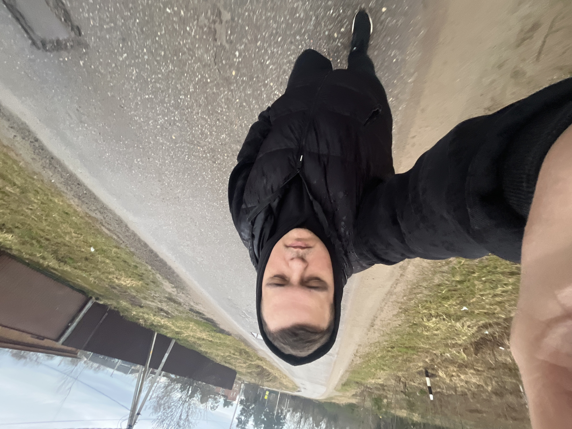 A man is standing on a street in Раздолье, Russia. He is wearing a black hooded jacket and black pants, with black shoes visible in the background. The street is paved, with grass and vegetation visible on either side. In the background, there are buildings and trees. The image appears to be a selfie taken at an angle.