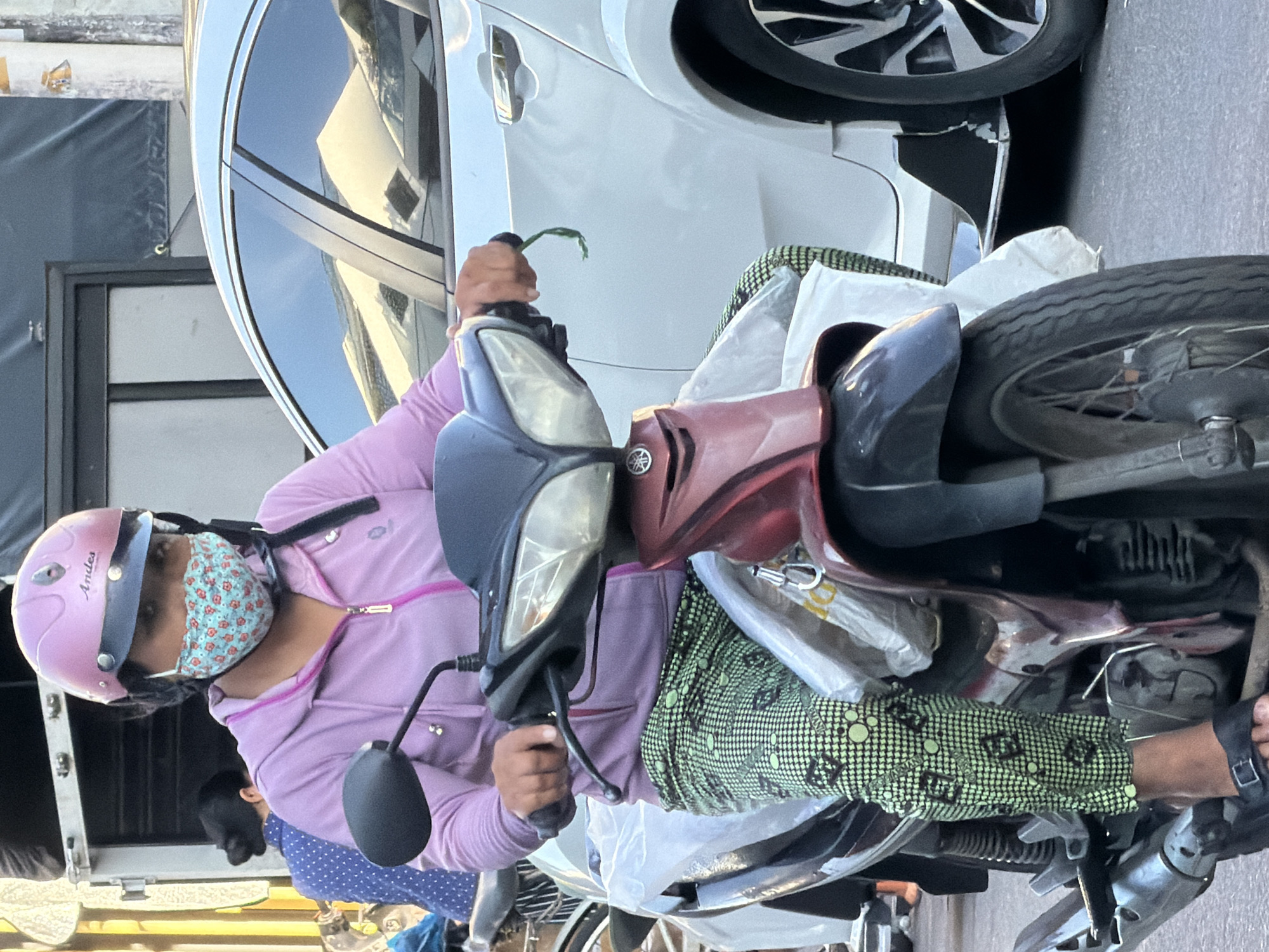A person wearing a pink helmet and a floral-patterned face mask rides a motorbike in Nha Trang, Vietnam. The rider is wearing a light purple hooded jacket and green patterned pants. A white car is parked next to the motorbike. The scene appears to be on a street in the city.
