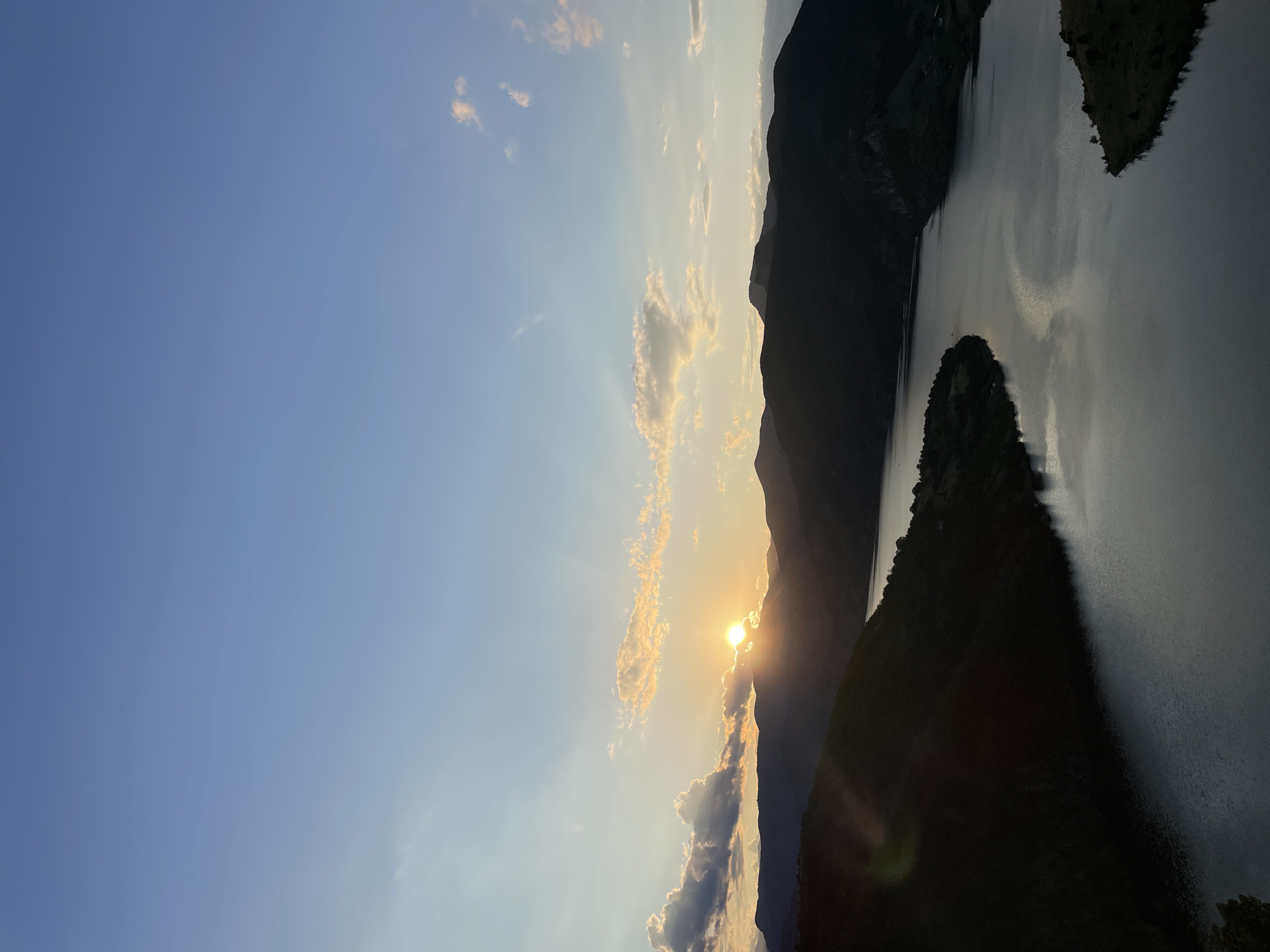 An aerial, wide-angle shot captures a serene sunset over a vast, winding river nestled between silhouetted mountains in Borovitsa, Bulgaria. The sun, a radiant orb, peeks from behind a dark mountain range, casting golden rays and illuminating wisps of scattered clouds. The tranquil river reflects the soft twilight hues of the sky. The foreground features a steep, shadowed incline covered in dark foliage. The overall scene is one of natural beauty and peaceful solitude.