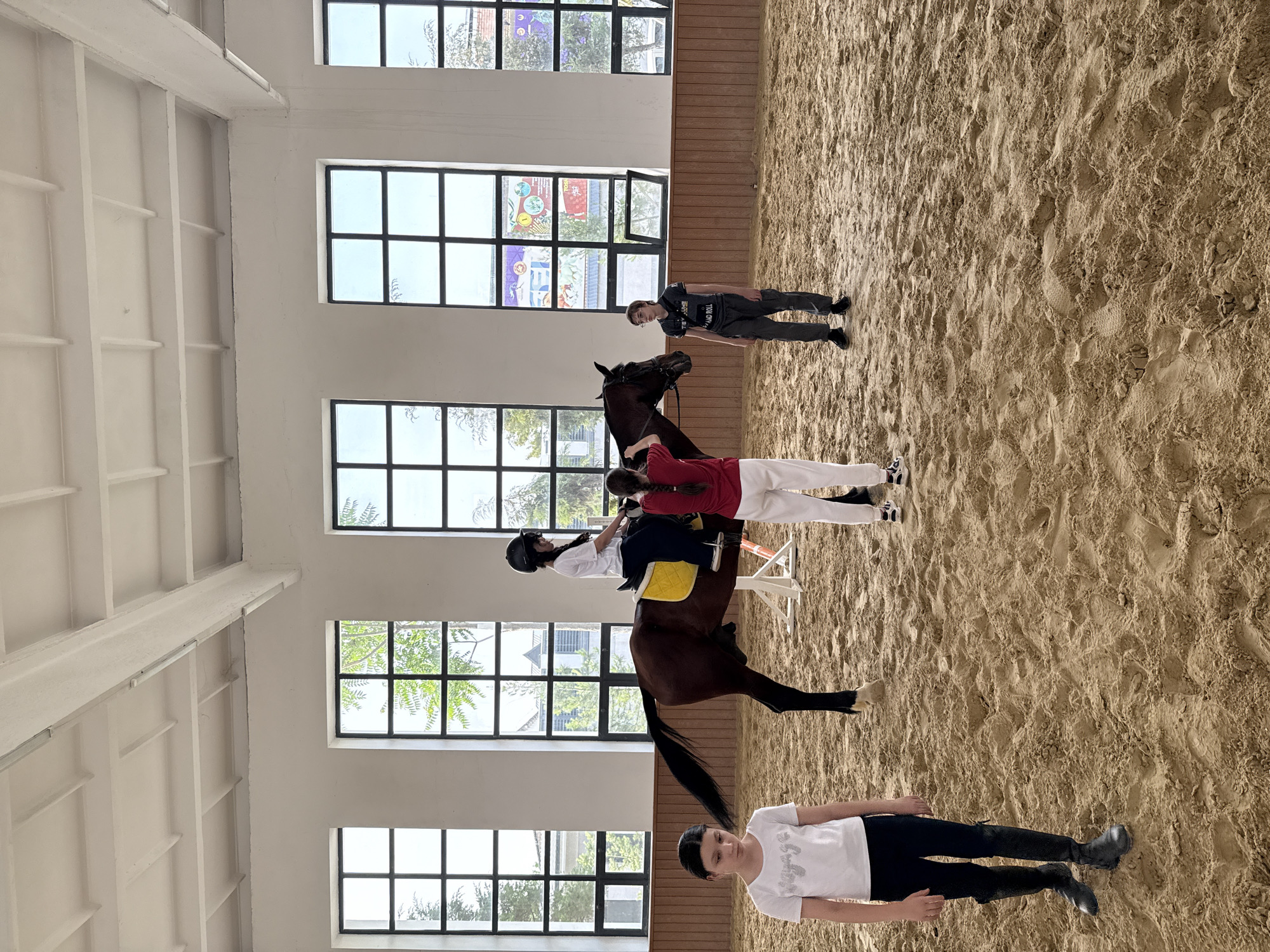 The image shows the interior of an equestrian arena. The arena has a sand floor and is illuminated by natural light through several windows on the left wall. Visible through the windows are trees and buildings, with one structure appearing to have colorful decorations.
A horse stands in the center of the arena, and there is a person wearing a red shirt and white pants standing behind it, presumably assisting the rider on horseback. The horse is dark brown, and the rider is wearing a white top, dark riding pants, and a helmet. They are seated on a yellow saddle pad. A woman in a white T-shirt and black leggings is on the ground in front of the horse. Another individual, wearing a dark top and pants, is standing farther in the background.
The location appears to be in Tashkent, Uzbekistan, due to the style of the building visible through the window which is similar to other buildings in the area.