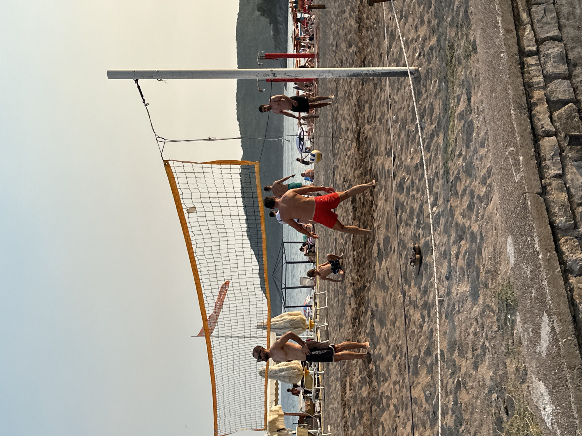 A group of men are playing beach volleyball on a sandy beach in Boreti, Montenegro. The sun is setting, casting a warm glow over the scene. The net is in the foreground, with several players visible on either side. In the background, there are umbrellas and lounge chairs set up on the beach, and a body of water with hills beyond. The players are wearing swim trunks and appear to be enjoying the game. The atmosphere is relaxed and casual.