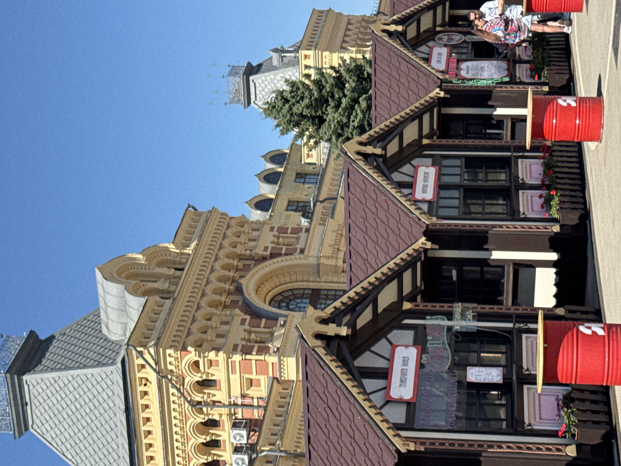 The image shows a street scene with two people standing near a building with a distinct architectural style. The building on the right has a dark-brown facade with a red tiled roof and decorative beams. It appears to be a series of small shops or kiosks, each with windows and signs. Two large red cylindrical objects, possibly barrels, are placed outside one of these units. The people are standing and looking towards the building. The person on the left is wearing a patterned shirt and shorts, while the person on the right is in a red and white plaid shirt and shorts. The building on the left is larger and has a more ornate facade with yellow and red brickwork, arched windows, and intricate decorative elements. A tall evergreen tree is visible between the two buildings, and a section of a grey tiled roof with a spire-like structure is also present. The sky is clear and blue, suggesting a sunny day. There is text visible on the red signs attached to the shops, but it is not clearly legible. The overall impression is of a marketplace or a tourist area with a mix of historical and contemporary elements. The scene appears to be outdoors during daylight hours, likely a warm, sunny day. The location context suggests Nizhny Novgorod, Russia.