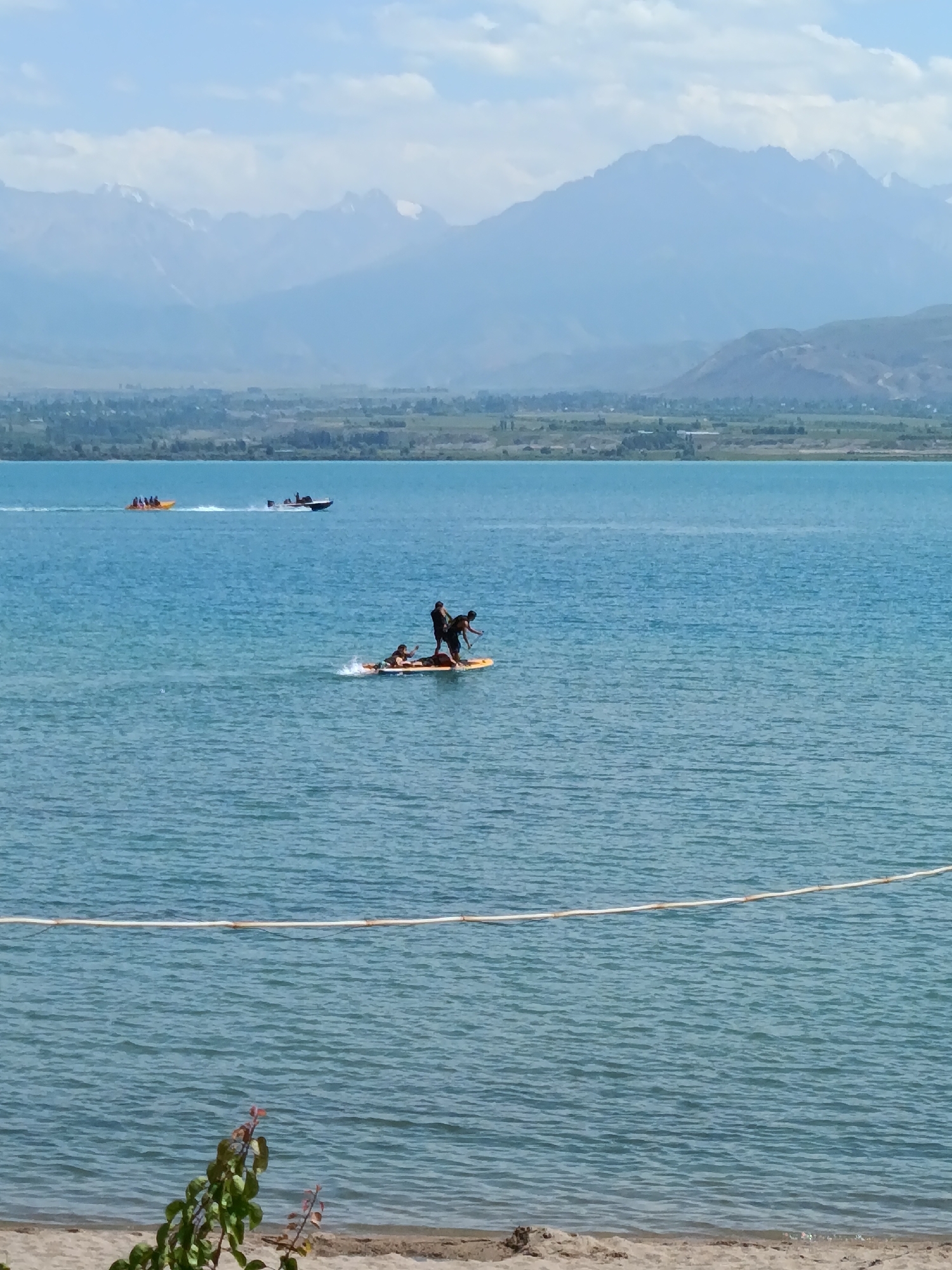 In Kyzyl-Suu, Kyrgyzstan, a panoramic view captures a serene lake with distant, snow-capped mountains under a hazy sky. The foreground features a sandy beach with a small branch of leaves. On the vibrant blue water, several recreational activities are underway. Two people are standing on a yellow paddleboard, one appearing to be instructing the other. Further out, a speed boat pulls a yellow inflatable banana boat carrying multiple passengers. A separate white speed boat is also visible in the distance. The water is calm with gentle ripples, and a white rope or buoy line is stretched horizontally across the middle ground, separating the swimming area from the open water.