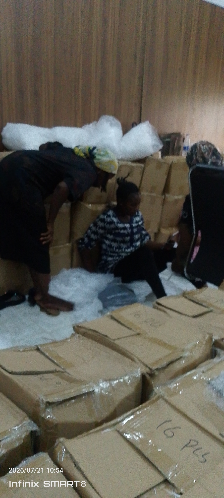 In Kakau, Nigeria, two women are seen in what appears to be a storage area. One woman, wearing a dark outfit and a headscarf, is bending over, looking down at some items. The other woman, dressed in a patterned top and dark trousers, is seated on the floor. They are surrounded by numerous cardboard boxes, some wrapped in clear plastic. The boxes are stacked and arranged on the floor, suggesting a warehousing or inventory setting. The environment features a wooden-paneled wall in the background and a tiled or concrete floor. The lighting is dim, and the overall atmosphere suggests a task involving the handling or organization of goods.