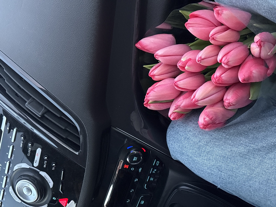 The image depicts the interior of a vehicle, viewed from a slightly elevated perspective. A person's lap, covered by light blue denim jeans, occupies the lower left and central portion of the frame. Resting on the lap is a dense bouquet of approximately 20-25 vibrant pink tulip buds, some with visible green foliage, wrapped in clear plastic or cellophane.
In the upper half of the image, the dark grey/black dashboard and central console of the vehicle are visible. The dashboard features a textured surface. Prominently displayed is a car stereo and climate control panel. The upper section of the console includes a main rotary knob, labeled "OK" in its center, surrounded by buttons such as "DISPLAY," "SOUND," "MUTE," "INFO," "CLOCK," "TA," and a red triangular hazard light button. Below this, a secondary control panel features additional rotary knobs, including one marked "MAX A/C," and various other climate and vehicle function buttons. Black air vents are visible to the right of the upper control panel.
The specific location within the city of Slavyansk-na-Kubani, Russia, is not discernible from the visual information presented in the image.