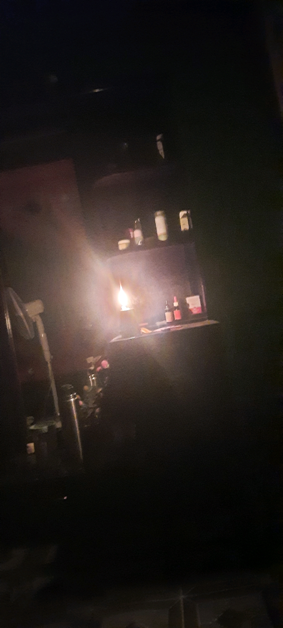 The image depicts a dimly lit interior, possibly a room or a shop, with a prominent oil lamp burning, casting a warm glow and creating atmospheric haze. The lamp is positioned on a surface, and to its right, a shelf displays several bottles and containers. In the foreground to the left, a metallic thermos or flask is visible, along with some pink flowers. A white fan is also partially visible in the lower left corner. The overall impression is one of a rustic or traditional setting, illuminated by the soft light of the oil lamp. The location in the city of Khongjom, India, adds a cultural context to the scene, suggesting it might be a representation of local life or a specific establishment within the city.