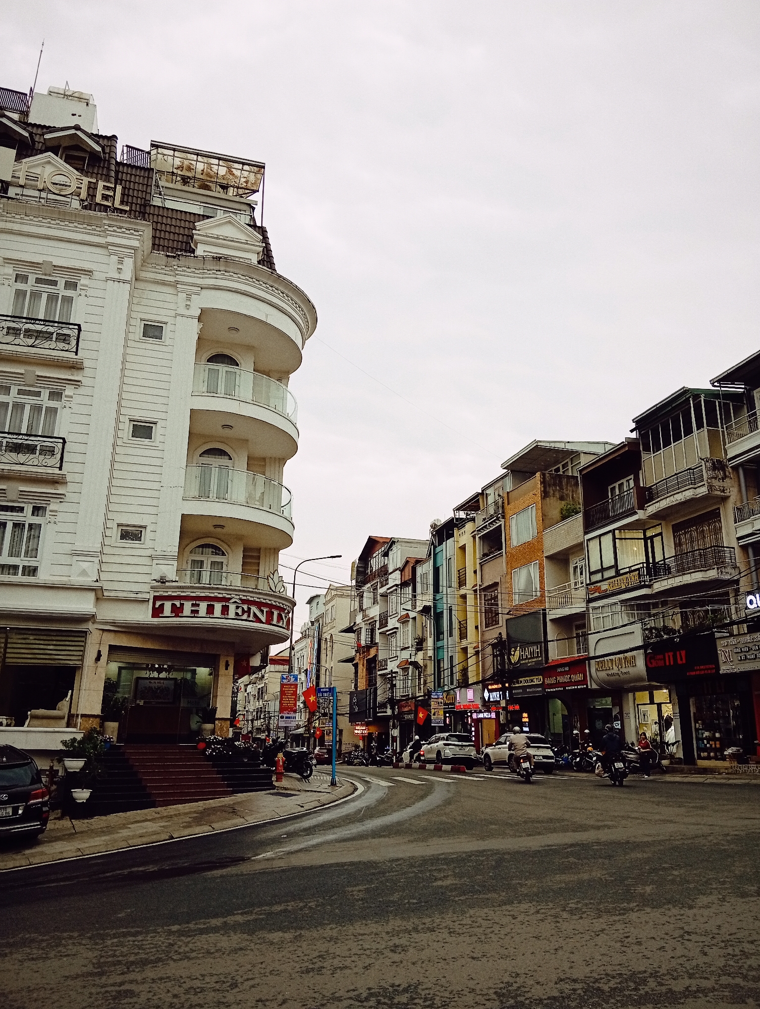 This media file captures a bustling street scene in Da Lat, Vietnam, under an overcast sky. The image features a prominent, elegant white hotel building on the left, identified by the large "HOTEL" sign on its roof and "THIEN LY" sign over its entrance. This building boasts multiple balconies with decorative railings and a curved facade.
The street, appearing somewhat damp, curves to the right, lined with a continuous row of multi-story buildings, each with distinctive balconies and ground-floor businesses. These buildings display a variety of architectural styles and colors, ranging from muted tones to brighter hues like orange and yellow.
The activity on the street depicts daily urban life. Several motorbikes, a common mode of transport in Vietnam, are visible; some are parked, while others, carrying riders, are in motion along the road. A black SUV is parked near the hotel, and a white sedan is further down the street. People can be seen near the shops and on motorbikes.
Notable details include various street signs and advertisements in Vietnamese, confirming the location. Visible text from the shops includes "HAIYIH," "JUNG HO," "KELLY QUYNH Wedding Event," and "BÀ TRIỆU" (likely a street name). Other partial texts mention "MOC LONG TEA," "ĐẶNG PHƯỚC QUÂN," "TƯ VẤN - THIẾT KẾ" (Consultation - Design), and "LÀNG PIZZA" (Pizza Village). The general impression is one of a lively commercial district during daylight hours on a cloudy day.
