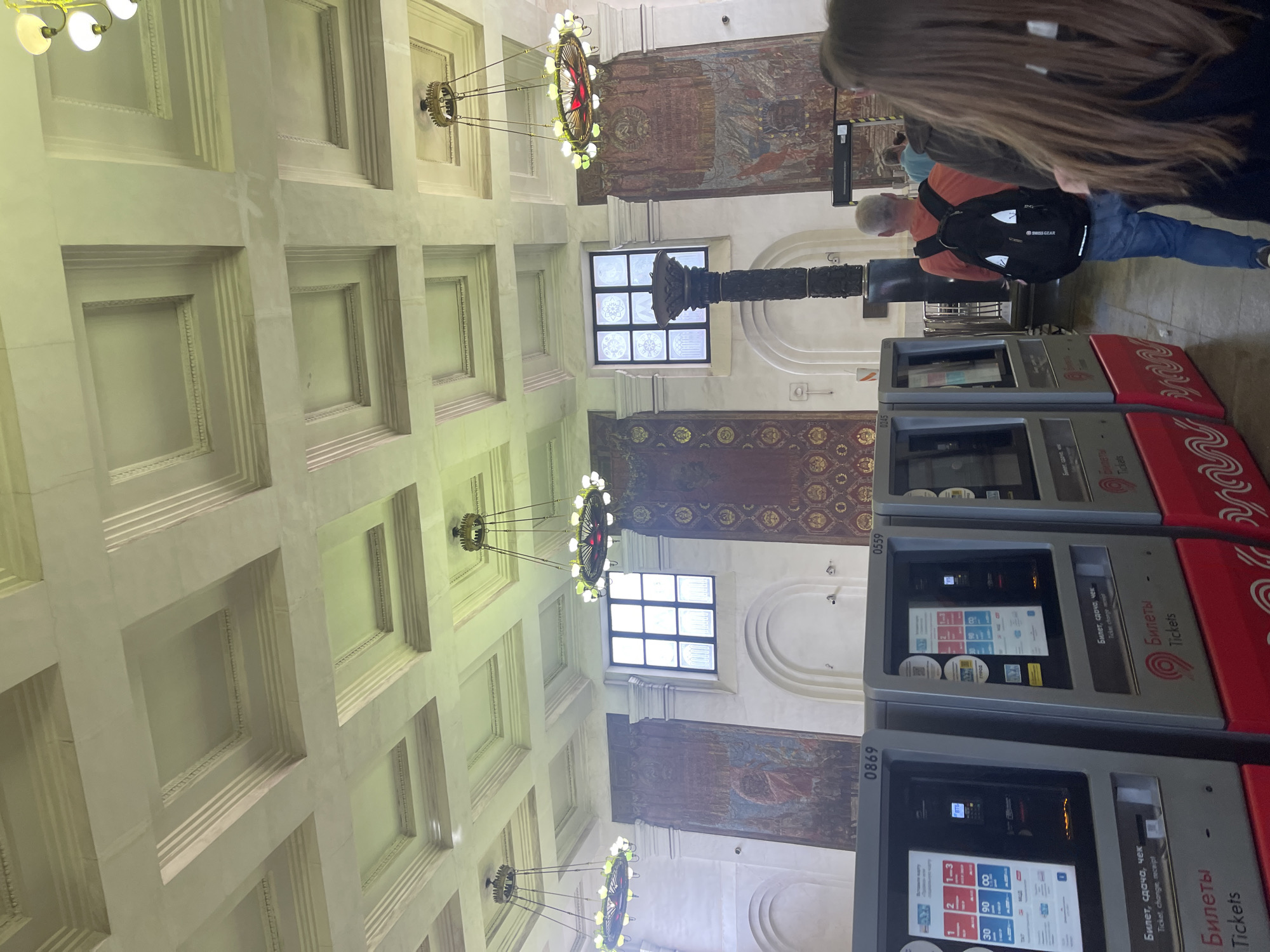 The image depicts the interior of a grand hall, likely a train station in Moscow, Russia, characterized by its high, ornate ceiling with a coffered design and large, multi-tiered chandeliers. Several ticket vending machines are visible in the foreground, with "Билеты Tickets" prominently displayed on their lower sections. The machines are labeled with numbers like 0869 and 0559. Large, decorative tapestries or murals adorn the walls between arched alcoves, featuring historical or symbolic imagery. One mural appears to incorporate the hammer and sickle symbol. A tall, dark, carved column stands near a window with a grid-like pattern. People are present, including a man with a black backpack and an orange shirt walking away from the camera, and the back of a woman with long brown hair in the foreground. The environment appears clean and well-lit, suggesting an active public space.