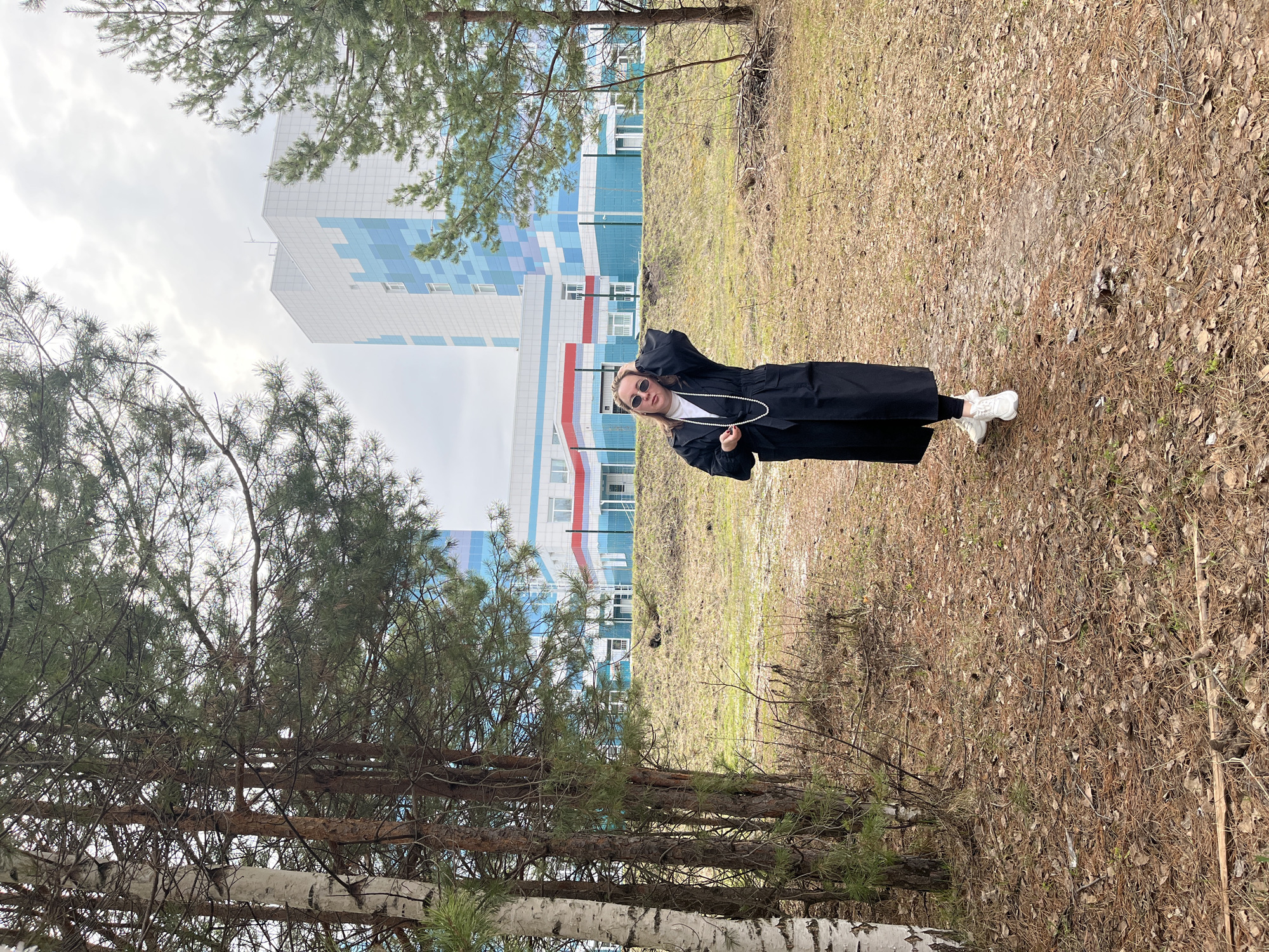 A woman stands on a grassy field with trees in Chaikovsky, Russia, near a tall building. She wears a black coat, white shoes, and sunglasses, and she is holding a pearl necklace. The ground is covered with dry leaves and vegetation. The building in the background is blue, white, and red. The sky is overcast.
