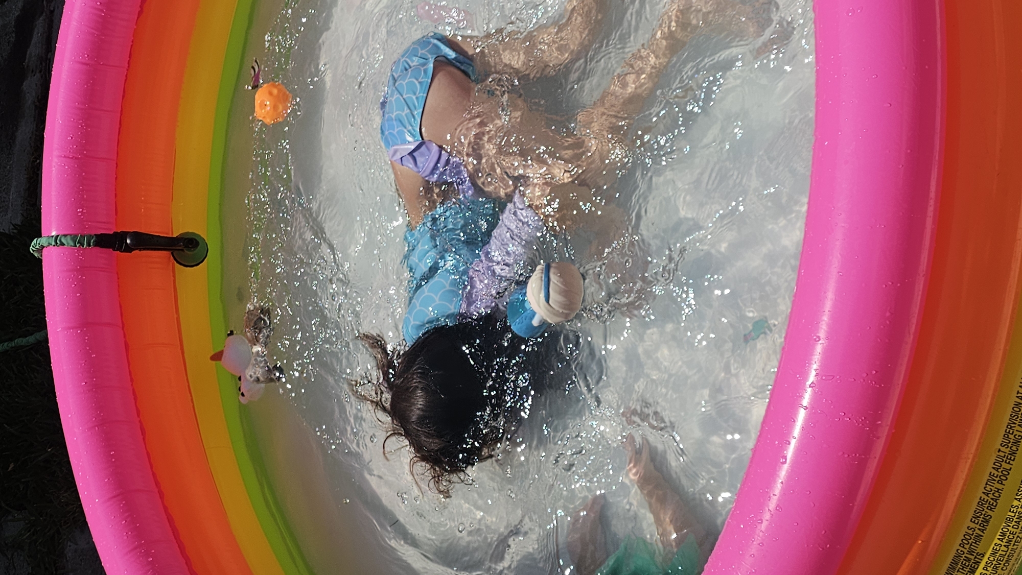 A child in a blue and purple swimsuit is partially submerged in a brightly colored, inflatable kiddie pool. The pool has pink, orange, yellow, and green rings. The child's face is not visible as they are partially underwater. Several plastic toys are floating in the pool. Part of a black suction cup toy is affixed to the pink pool wall. The pool is set outdoors on a patch of grass. The edge of the pool displays safety warnings in English and French. The location is San Bernardino Tlaxcalancingo, Mexico.