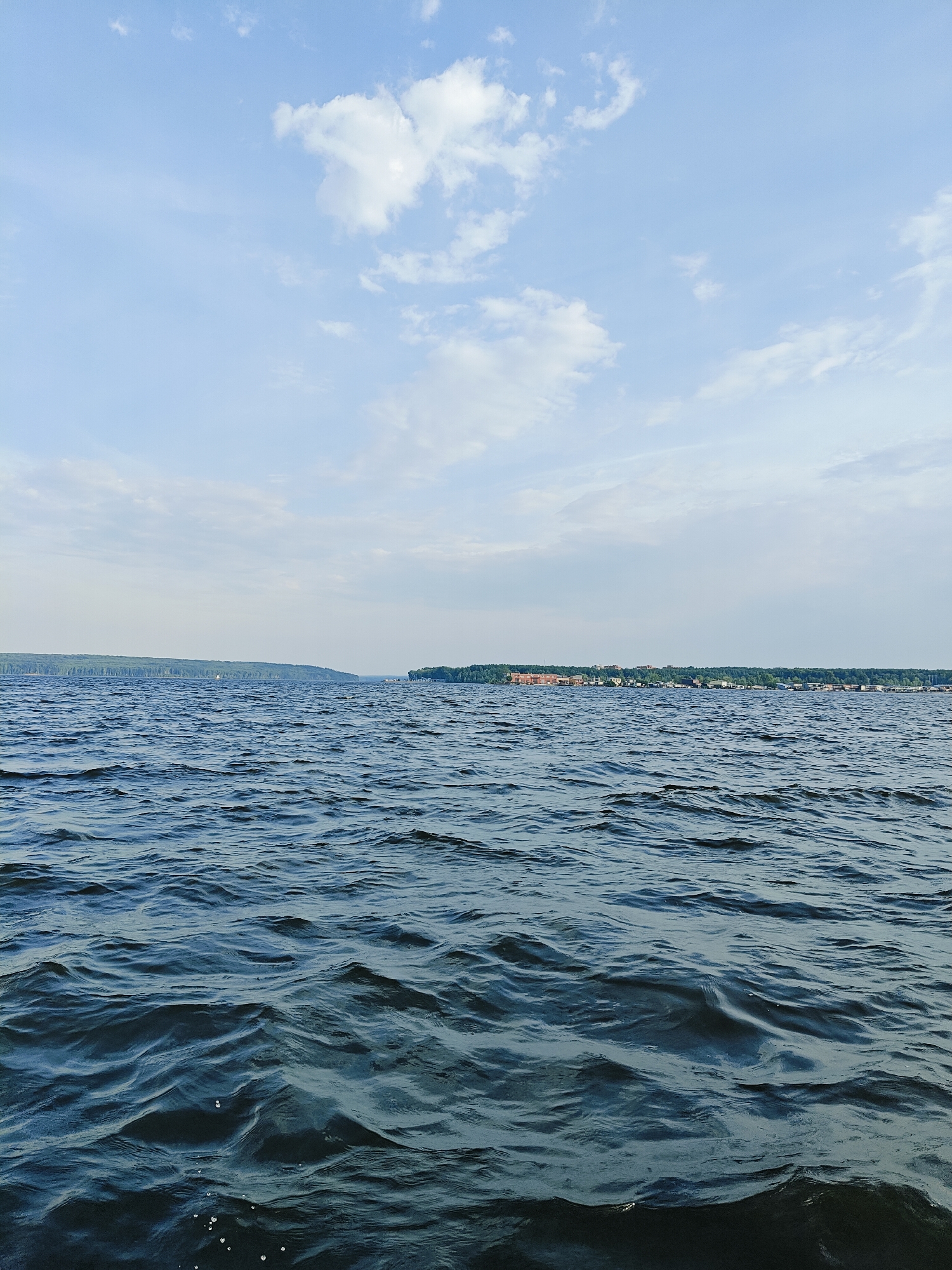 The image depicts a wide expanse of dark blue water with moderate waves under a bright, pale blue sky with scattered white clouds. In the distance, a long, tree-covered shoreline stretches across the horizon. On the right side of the shoreline, a settlement with red-tiled roofs and various buildings is visible, interspersed with green trees. Further to the left, a darker, denser forest lines the coast. A small white sailboat is visible in the distance on the water, to the left of the settlement. The overall scene suggests a serene, natural environment, likely a large river or lake, with human habitation present along the distant shore. This view is consistent with the appearance of the Kama River near Chaikovsky, Russia, a city situated on its banks.