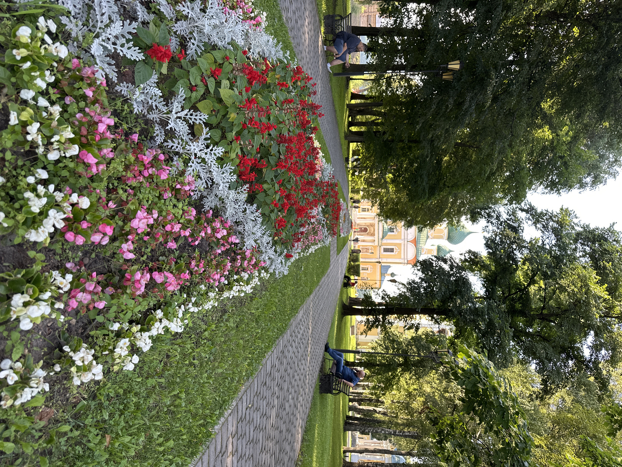 A paved walkway lined with flower beds runs through a park in front of a yellow building with green domes. Tall green trees with dappled sunlight filtering through their leaves line the walkway. On a bench to the left, a person in a dark shirt and blue shorts sits facing away from the camera, looking at their phone. On a bench to the right, a person in a blue shirt and dark pants sits facing the camera, also looking at their phone. The flower beds contain red, pink, white, and silver foliage plants. The sky is bright, indicating daytime.
