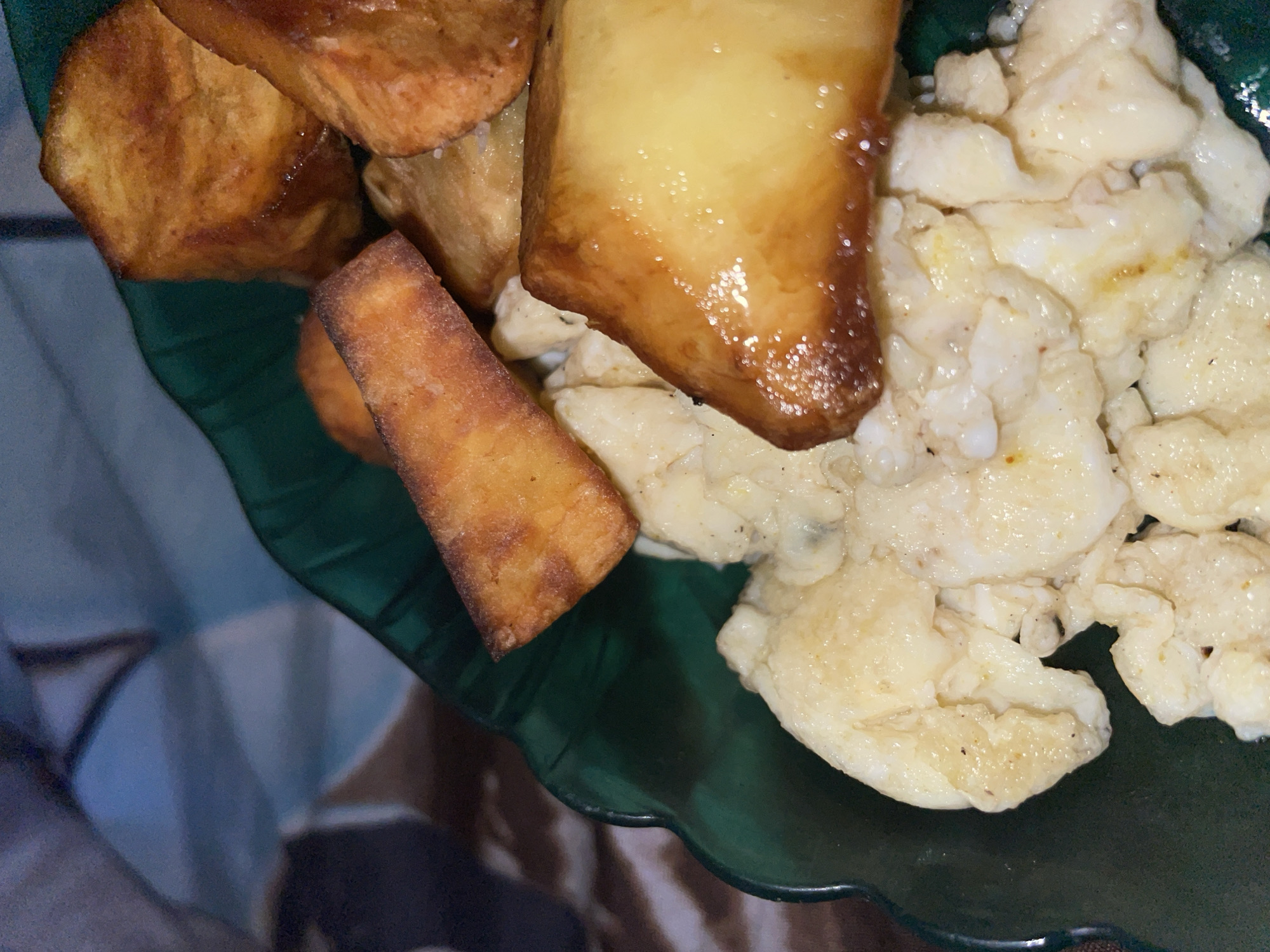 The image shows a close-up of food served in a green, wavy-edged bowl. The food appears to be scrambled eggs and roasted potatoes. The potatoes are cut into rustic, irregular shapes and are golden brown with some glistening oil. The scrambled eggs are light yellow and appear fluffy. The background is blurred but suggests an indoor setting, possibly a dining table, with a patterned surface visible. The image does not provide any information about the location in Barako, Nigeria.