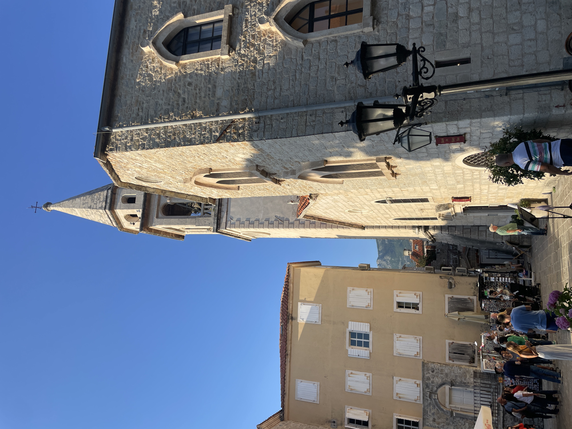 In Budva, Montenegro, a narrow street is illuminated by the warm glow of the sun under a clear blue sky. A prominent stone church with a tall, pointed steeple dominates the right side of the frame. The church's facade features arched windows and is constructed from light-colored stone blocks. To the left, a contrasting building with a yellow facade and white shutters stands. Several people are scattered throughout the scene, some browsing market stalls set up along the street, while others stand and converse. A vintage-style streetlamp with two lanterns stands in the foreground, casting shadows on the pavement. The overall atmosphere is one of a vibrant, sun-drenched European street scene.