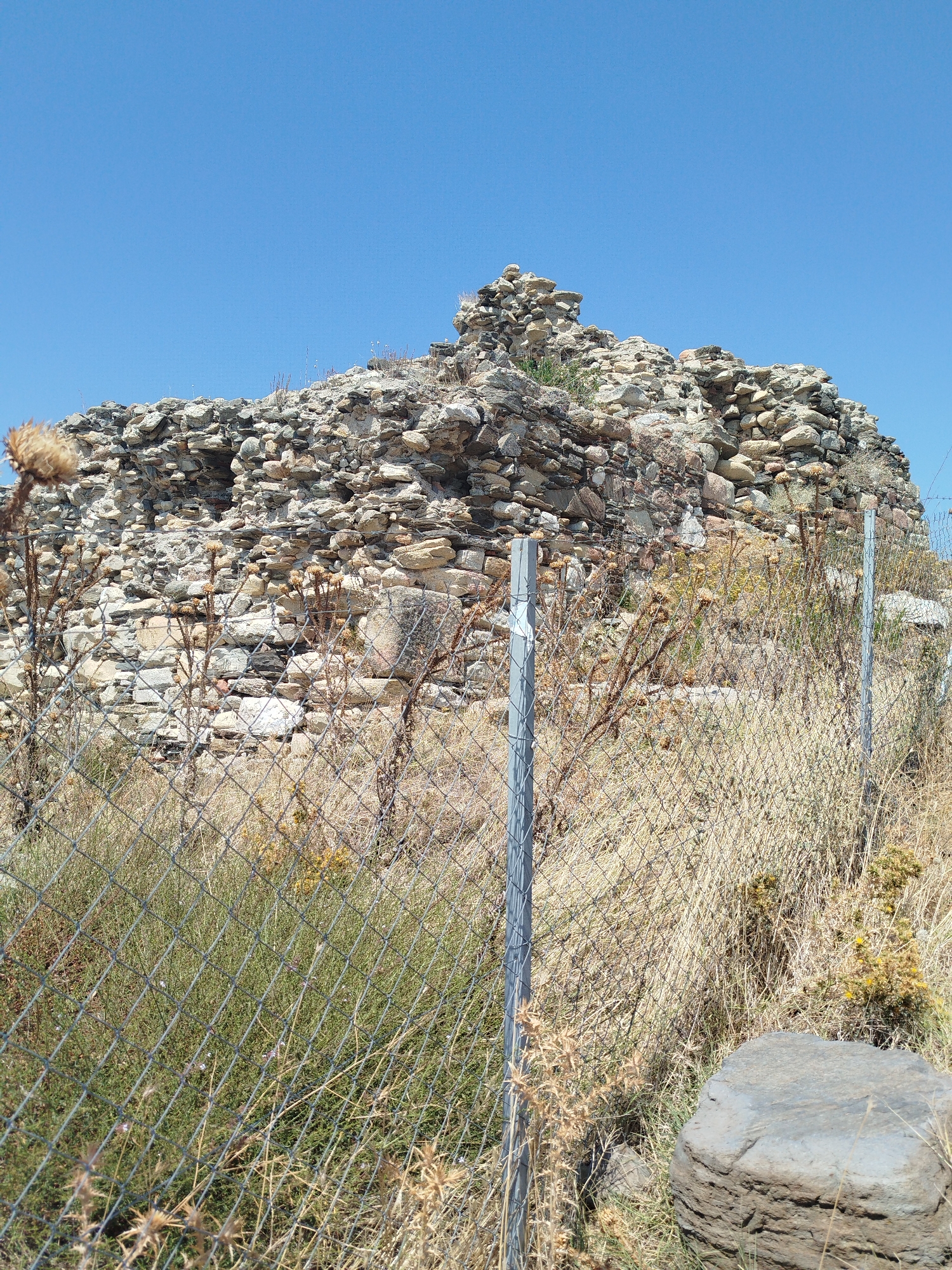 The image depicts a portion of ancient ruins located in the Sithonia Municipal Unit, Greece. The ruins are constructed from rough, irregular stones, forming a weathered and crumbling structure. In the foreground, a chain-link fence runs across the lower part of the frame, obscuring some of the lower ruins and surrounding vegetation. The vegetation consists of dry, brown grasses and some green bushes, interspersed with thorny plants. A large, rough-textured rock sits in the lower right corner of the image. The sky above is a clear, bright blue. There are no people or animals visible in the scene. The overall impression is one of an archeological site in a rural, somewhat arid environment.