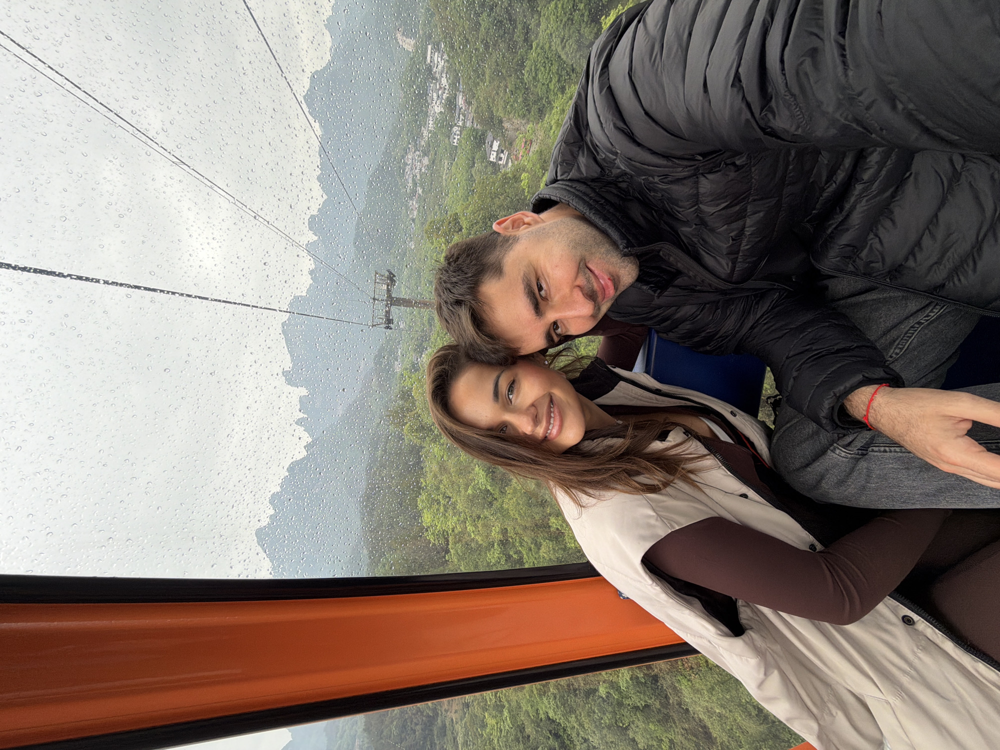 A couple is in a cable car overlooking a forested landscape near Zhangjiajie, China. The man is wearing a black puffer jacket and gray jeans. The woman is wearing a white coat over a brown shirt. Raindrops cover the window. The cable car is orange and black. The backdrop shows misty mountains and a town.