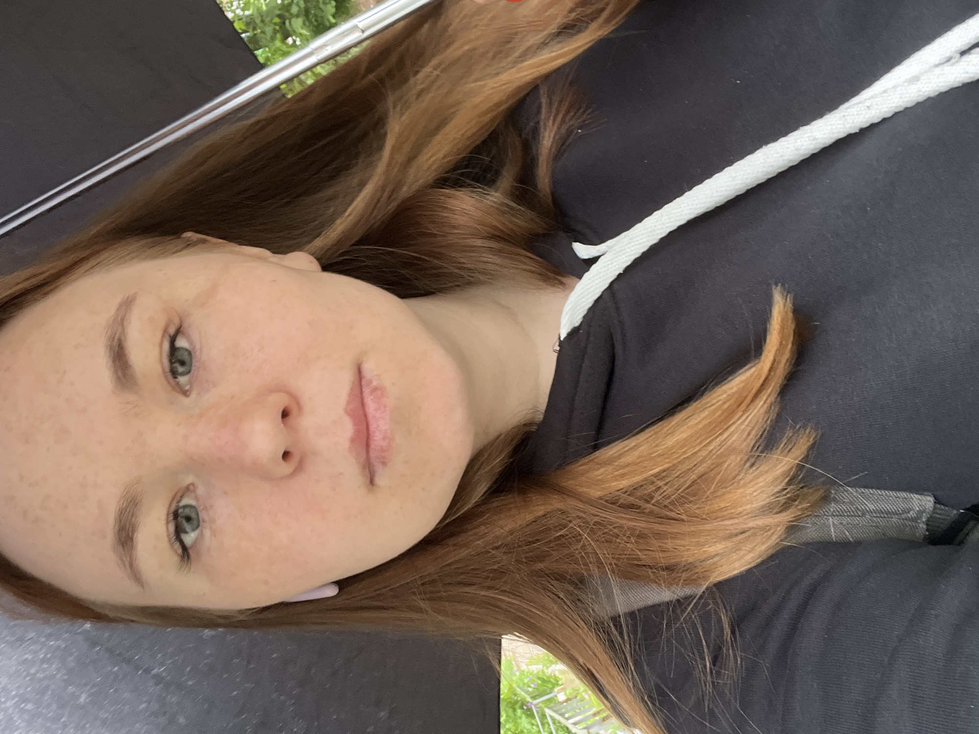 A close-up, diagonal portrait features a young woman, likely in Chaikovsky, Russia. Her head is positioned towards the bottom left, and her body extends diagonally upwards to the right. She has fair skin dotted with numerous freckles across her nose and cheeks. Her eyes are a light shade, appearing green or blue, and she gazes directly at the viewer with a neutral, calm expression. Her long, reddish-brown hair flows around her head and shoulders.
She is wearing a black hooded sweatshirt, partially visible, characterized by a prominent white drawstring around the hood opening. The background is mostly indistinct. In the upper left and upper right sections, a dark, possibly fabric-like surface (like an umbrella or canopy) is visible, bordered by a thin, reflective metallic bar. Beyond this, faint glimpses of green foliage and a light-colored structure suggest an outdoor or partially covered outdoor setting. The lighting is soft and natural, indicating daytime. There is no visible text within the image.