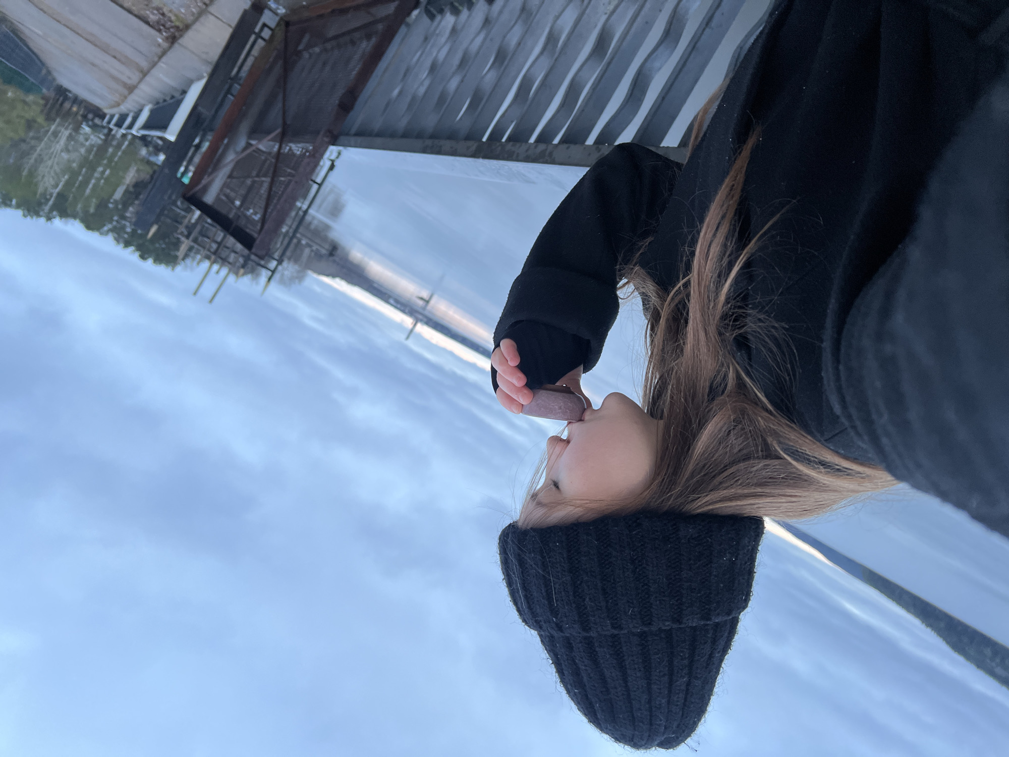 A young person wearing a black beanie and a black coat is depicted outdoors in a vertical, medium-shot composition. The person is holding and licking what appears to be a purple lollipop. The background consists of an overcast sky, a body of water that looks like a river or lake, and part of a metal structure resembling a pier or gangway. Based on the metadata, it is likely that the location is Chaikovsky, Russia, due to the date and time the image was taken, combined with the image content. The person is the main subject, and their action of licking the lollipop is the focal point of the image.