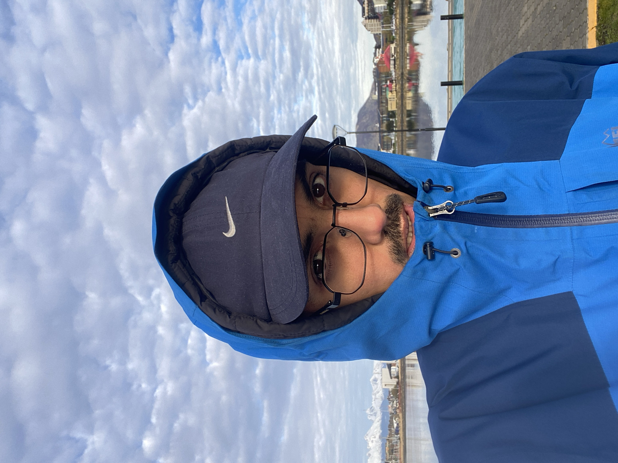 A selfie shows a man wearing a blue Nike baseball cap, glasses, and a two-tone blue jacket with a hood, against a backdrop of cloudy sky and the city of Ushuaia, Argentina. Buildings and water are visible in the background.