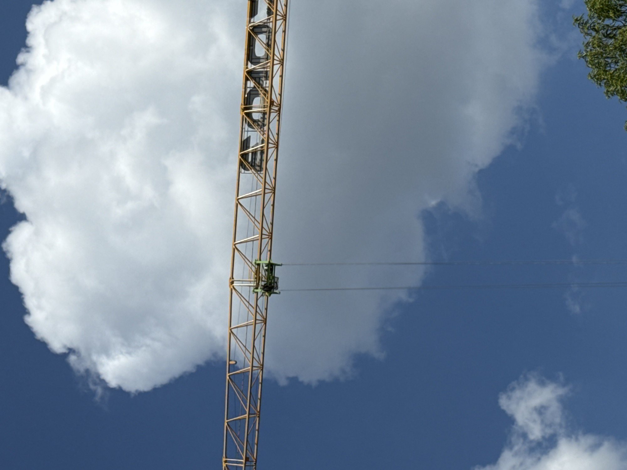 A construction crane's yellow lattice boom extends horizontally across a bright blue sky with large white cumulus clouds. A small, dark green and black mechanism is suspended from the underside of the boom by two thin wires. The crane structure has a visible logo or text that appears to be "EOCO". The image is taken from a low angle, looking up at the crane and the sky. The overall scene suggests an outdoor construction site on a clear, possibly sunny day.