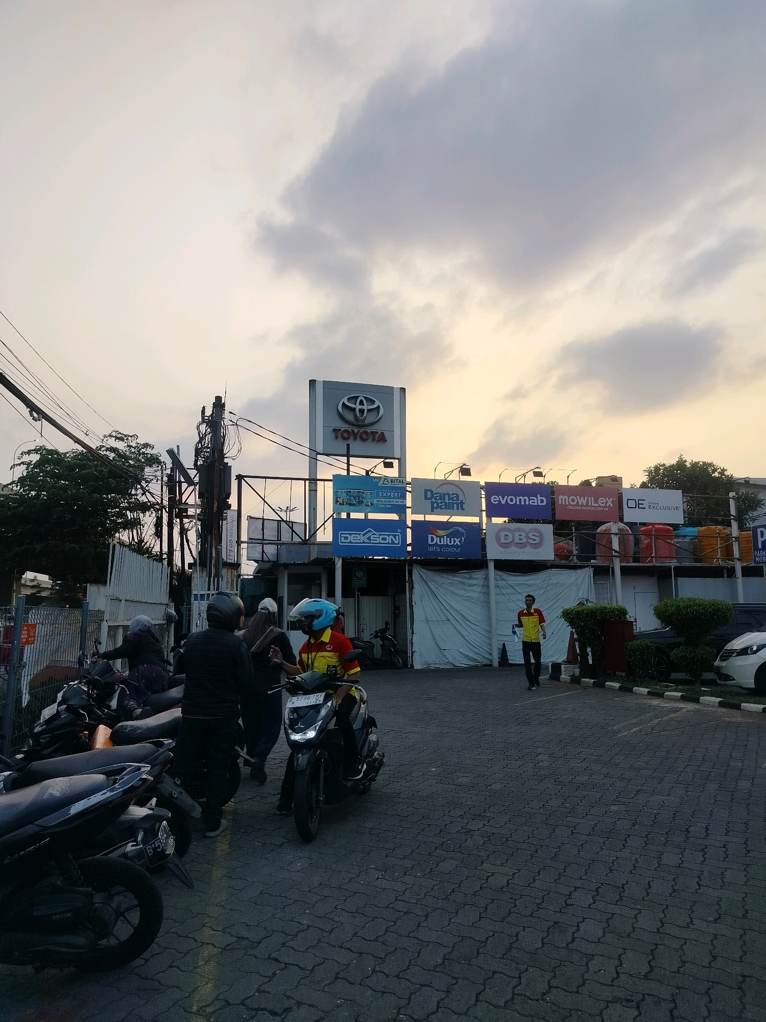 The image captures an exterior scene at a Toyota dealership in East Jakarta, Indonesia, likely during the late afternoon or early evening due to the sky's lighting. The dealership's prominent sign with the Toyota logo is visible. Adjacent to it, a series of large billboards advertise various brands, including Dana Paint, Evomab, Mowilex, OE Exclusive, Dekson, Dulux, and DBS. A person wearing a yellow and red shirt is walking in front of the building. Several motorcycles are parked in the foreground, and a few individuals are standing or sitting near them. The ground is paved with interlocking bricks. The overall impression is that of a commercial area with an automotive dealership and associated businesses.